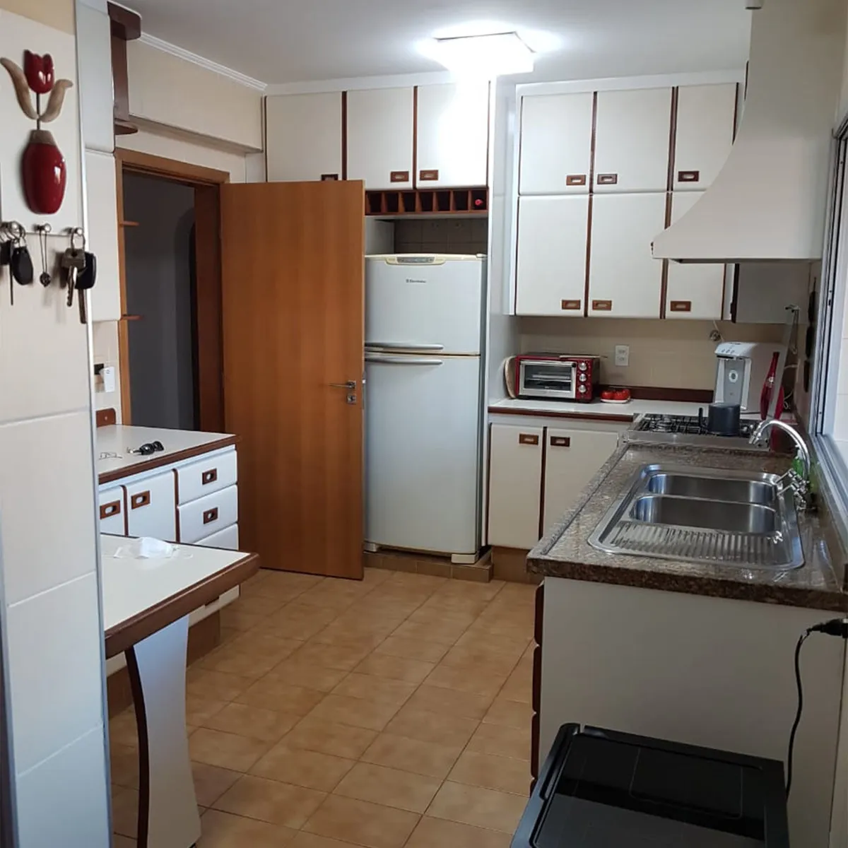 Cozinha antiga e estreita, com armários brancos de laminado, piso de cerâmica bege e coifa antiga, antes da reforma