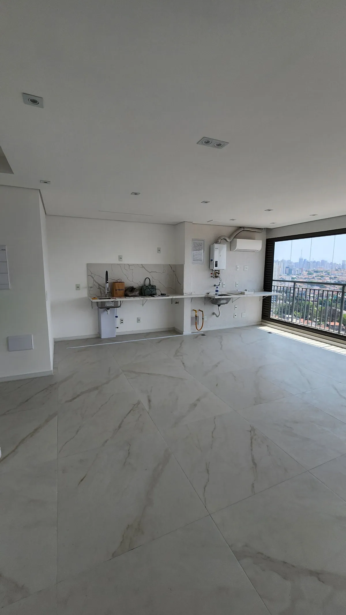 Sala ampla vazia com piso de porcelanato marmorizado, bancada com pia, aquecedor a gás e varanda envidraçada, antes da reforma