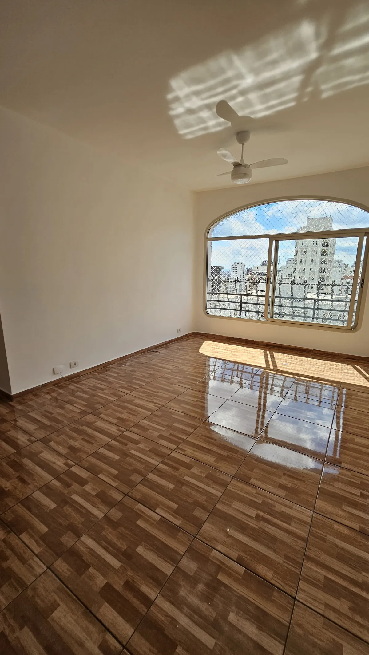 Sala vazia com piso de cerâmica marrom brilhante imitando madeira, ventilador de teto e janela em arco, antes da reforma