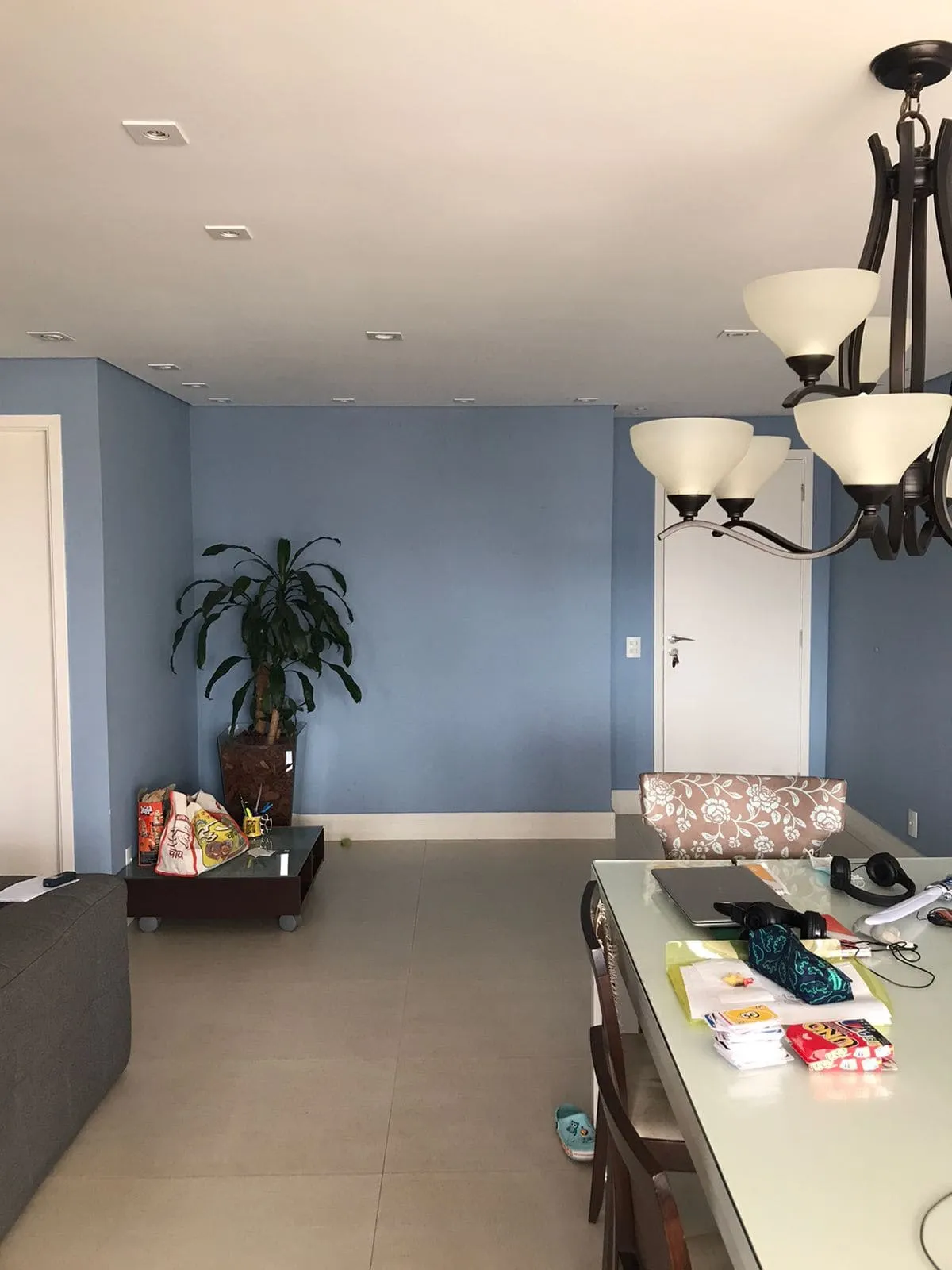 Sala com paredes azuis, lustre de ferro antigo, planta e mesa de vidro bagunçada, antes da reforma