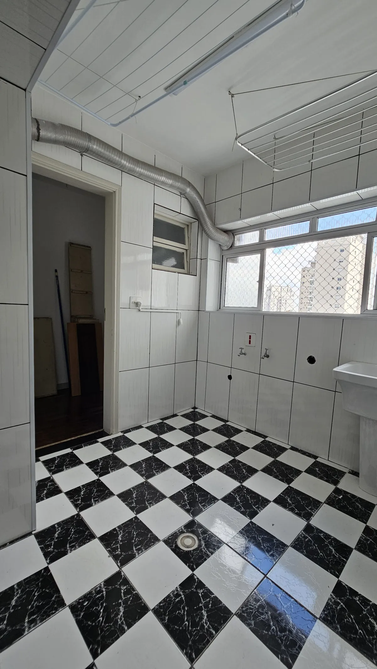 Área de serviço com piso xadrez, azulejo branco, varal de teto e janela para os prédios, antes da reforma