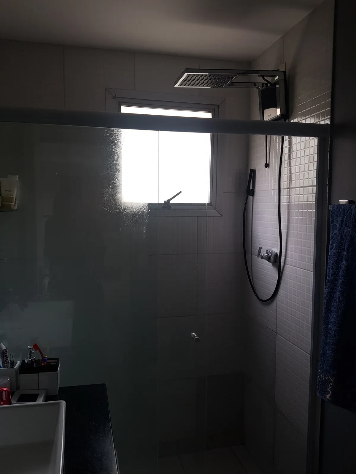 Box do banheiro com azulejo branco, ducha de teto, janela pequena e ambiente escuro, antes da reforma
