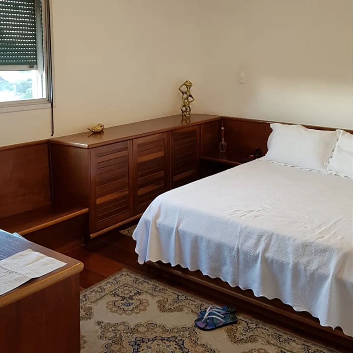 Quarto antigo com aparador de madeira escura, cama de casal e tapete estampado, antes da reforma