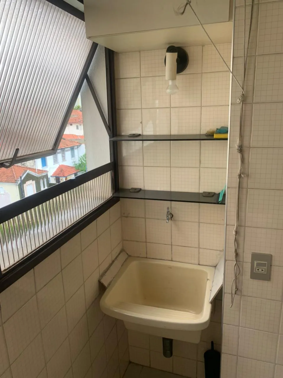 Área de serviço com tanque de louça, prateleiras de vidro e azulejo branco, janela para casas ao fundo, antes da reforma