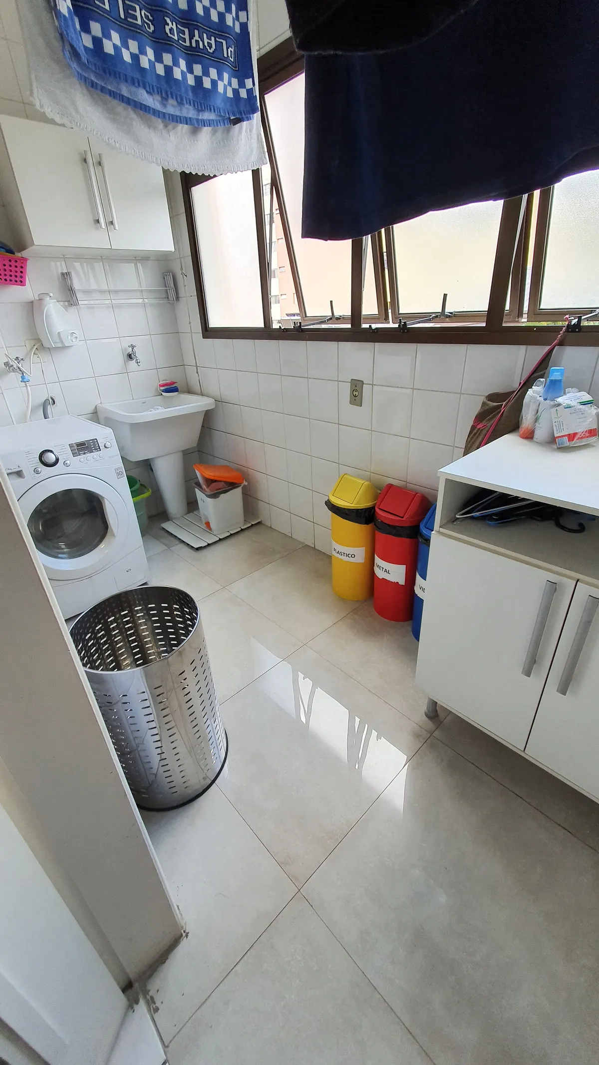 Área de serviço com máquina de lavar, tanque, azulejo branco e lixeiras de reciclagem, antes da reforma