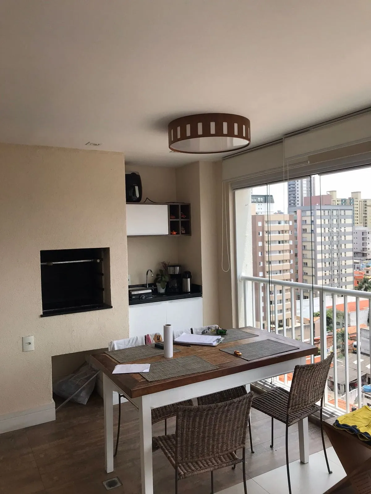 Varanda gourmet com churrasqueira, bancada branca, mesa de madeira e vista da cidade, antes da reforma