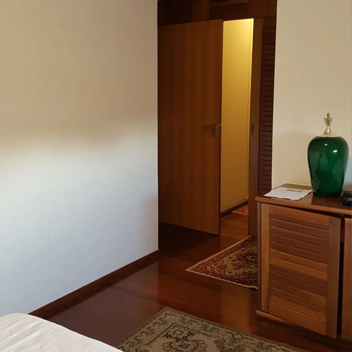 Quarto antigo com piso de madeira escura envernizada, porta de madeira e cômoda amadeirada, antes da reforma