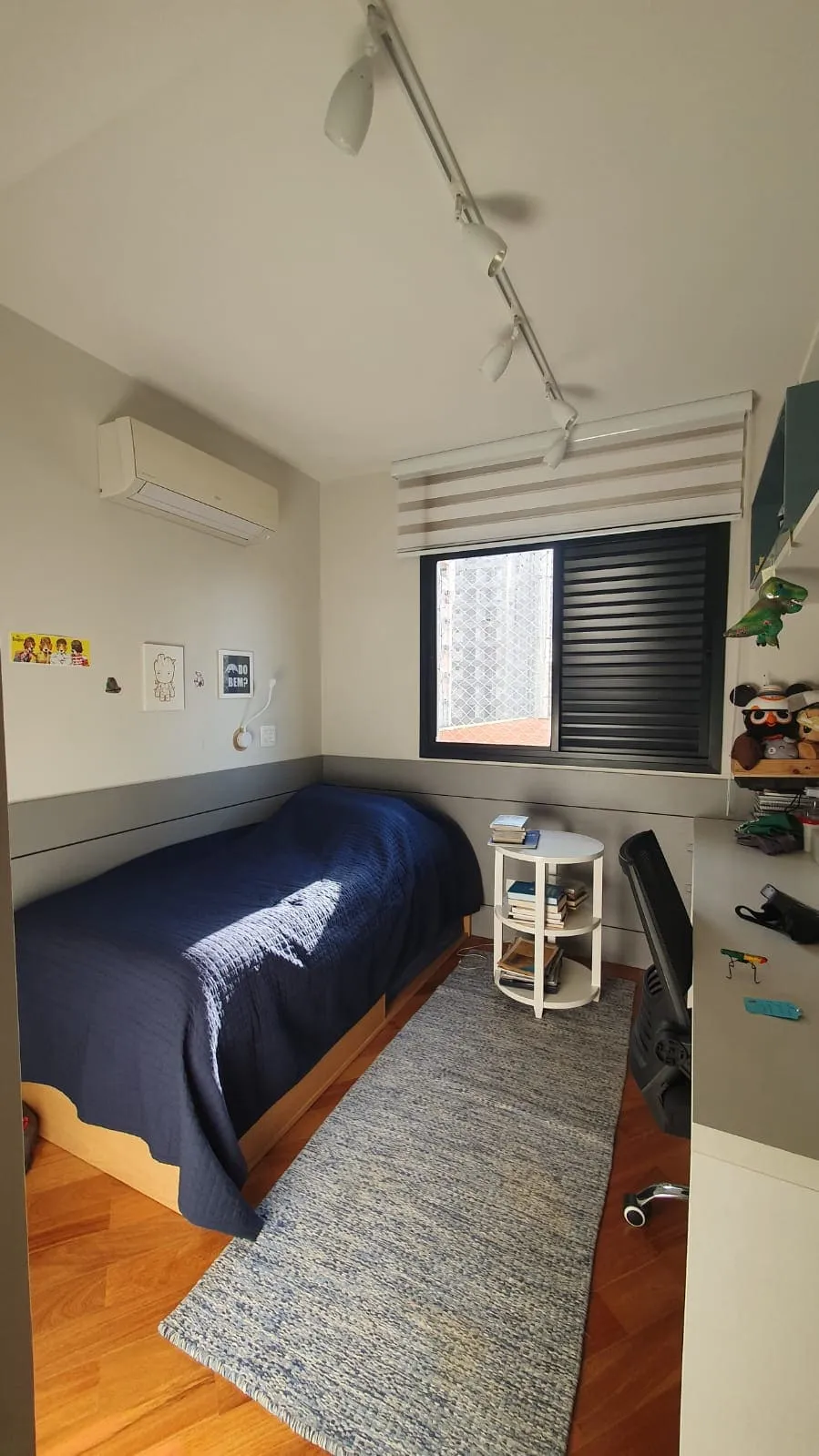 Quarto com cama de colcha azul, tapete azul trançado, mesa lateral branca e piso de taco, antes da reforma
