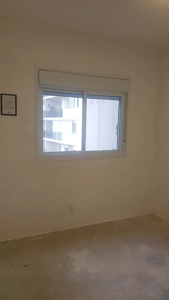 Quarto vazio de paredes claras com janela de correr e piso de contrapiso, antes da reforma
