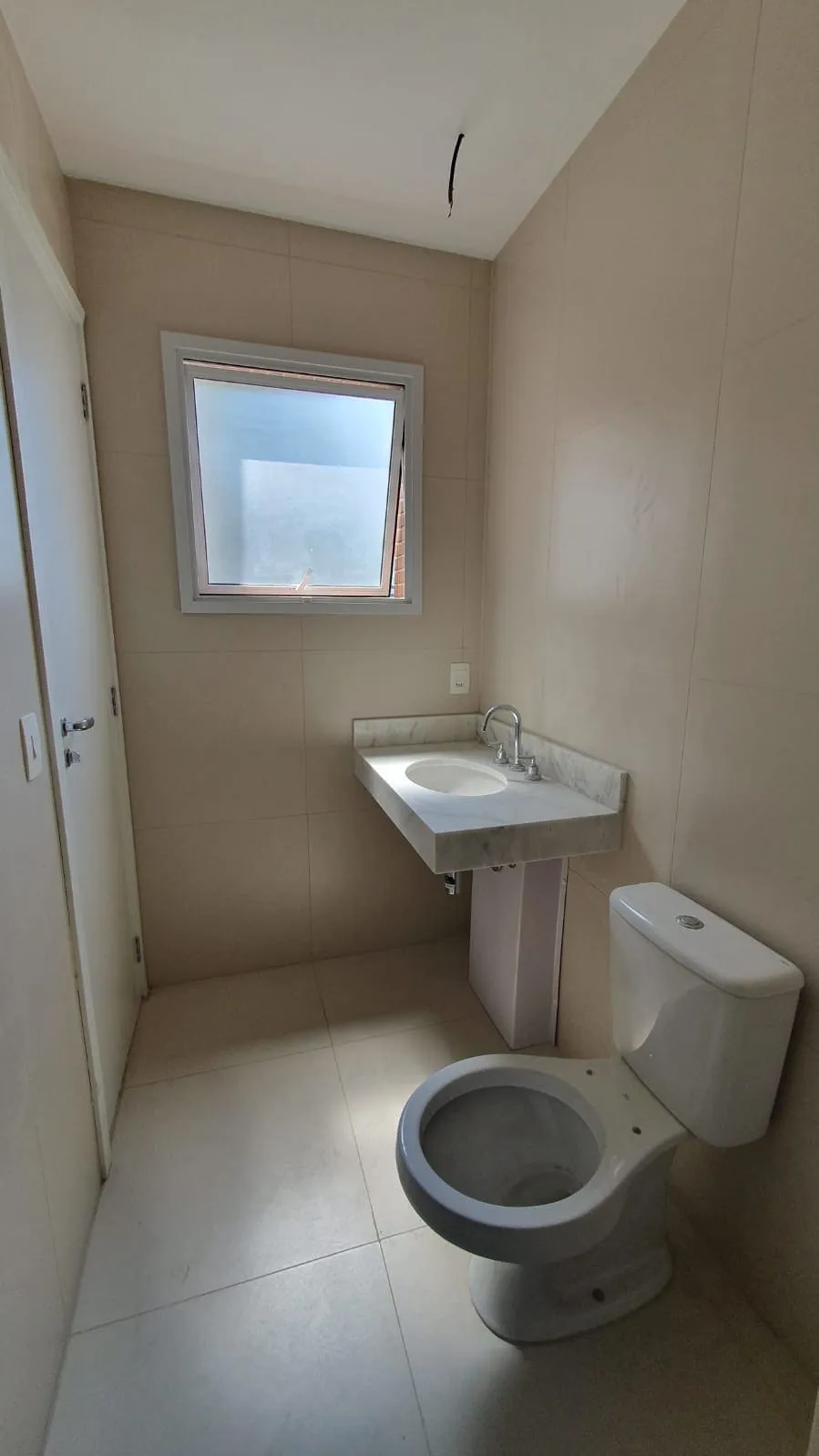 Banheiro com revestimento bege, bancada de mármore branco, vaso sanitário e janela basculante, antes da reforma