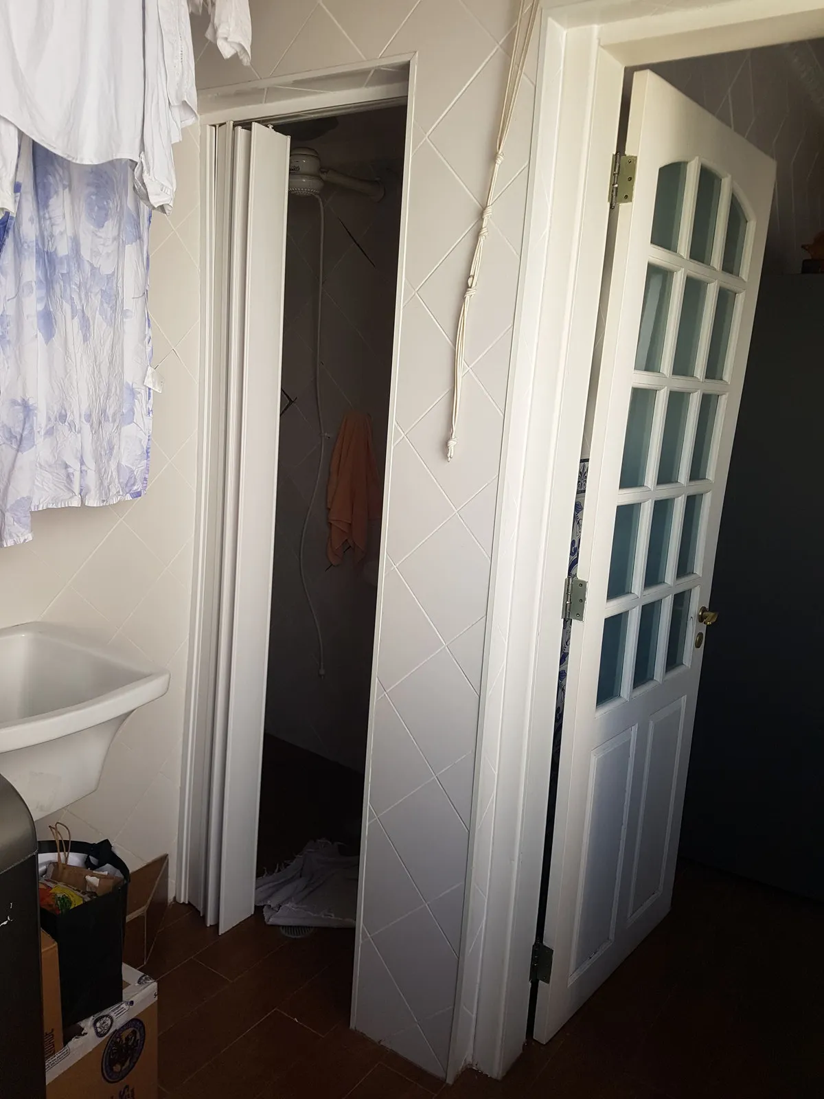 Banheiro antigo com porta sanfonada, box apertado revestido de azulejo e tanque branco, antes da reforma