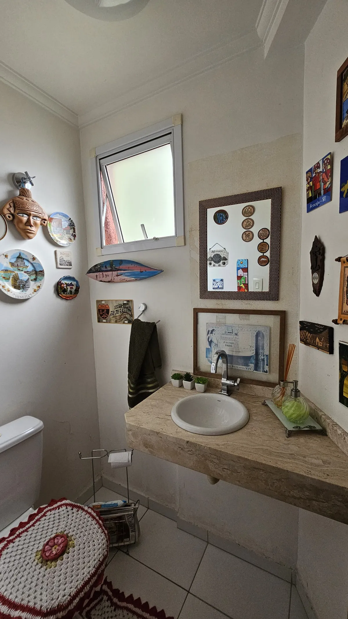 Lavabo antigo com bancada de mármore travertino, cuba de louça, espelho e parede cheia de pratos decorativos, antes da reforma