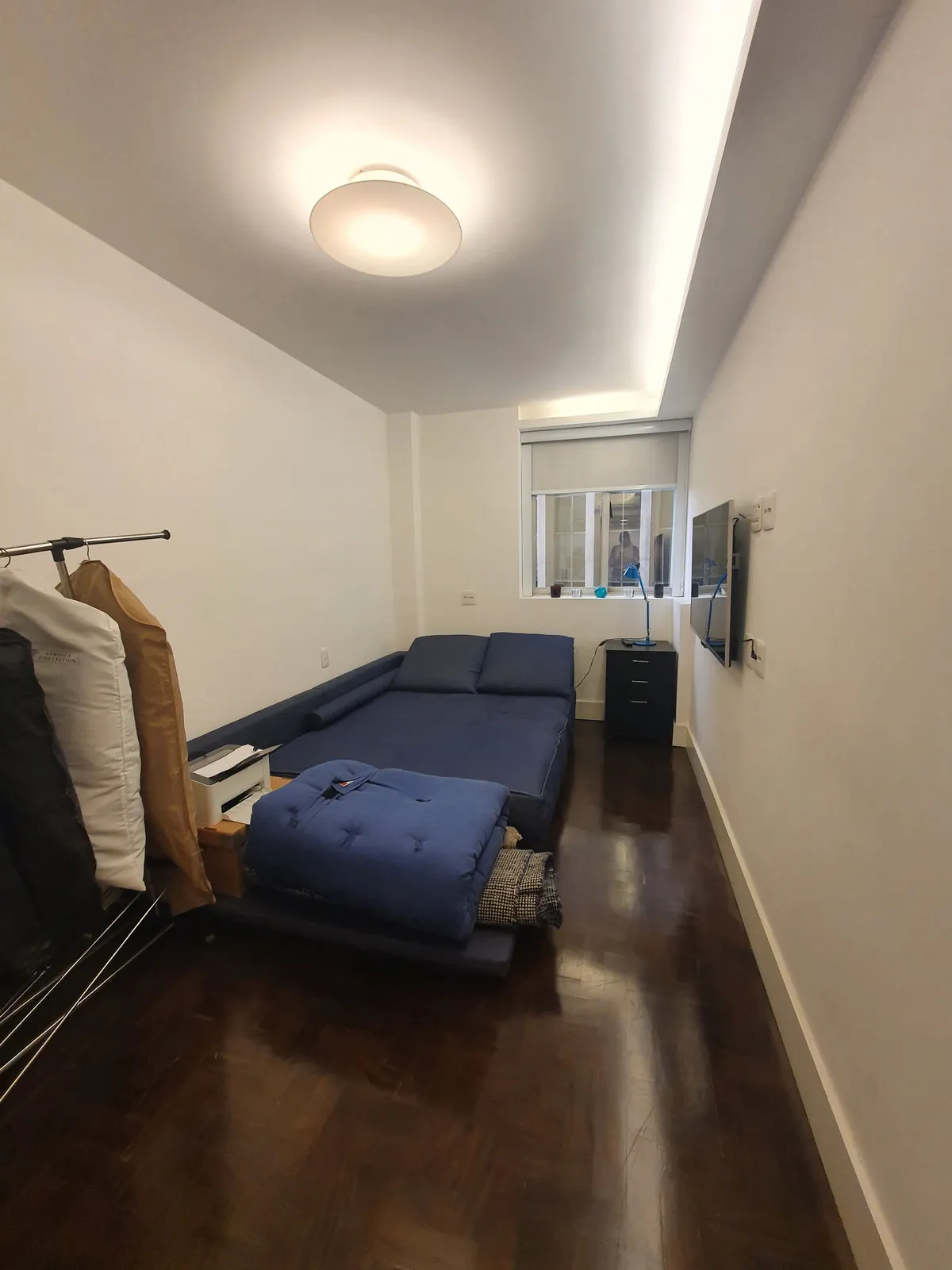 Quarto estreito com sofá-cama azul, arara de roupas e piso de madeira escura, antes da reforma