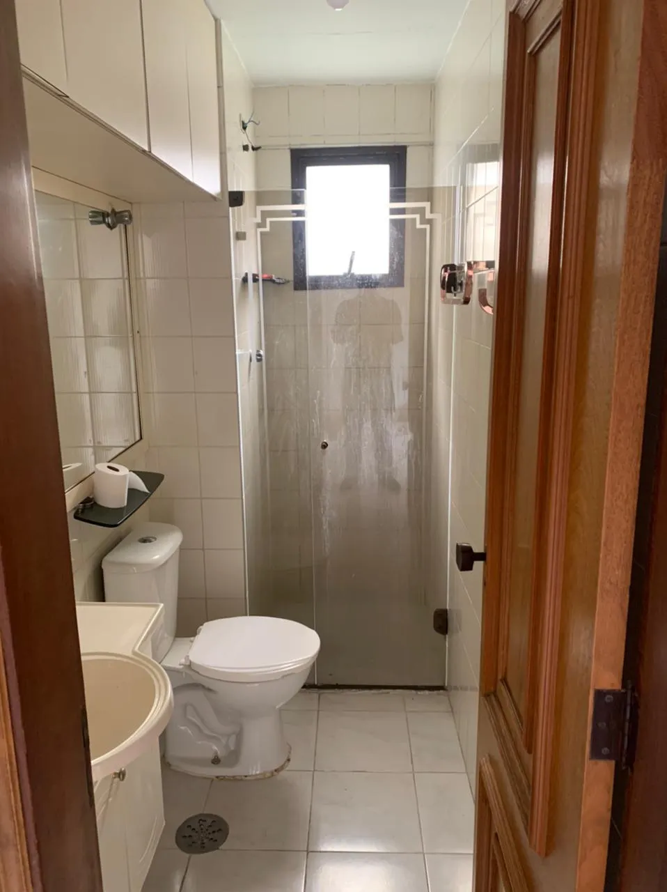 Banheiro estreito com azulejo branco, vaso sanitário, box de vidro jateado e porta de madeira, antes da reforma