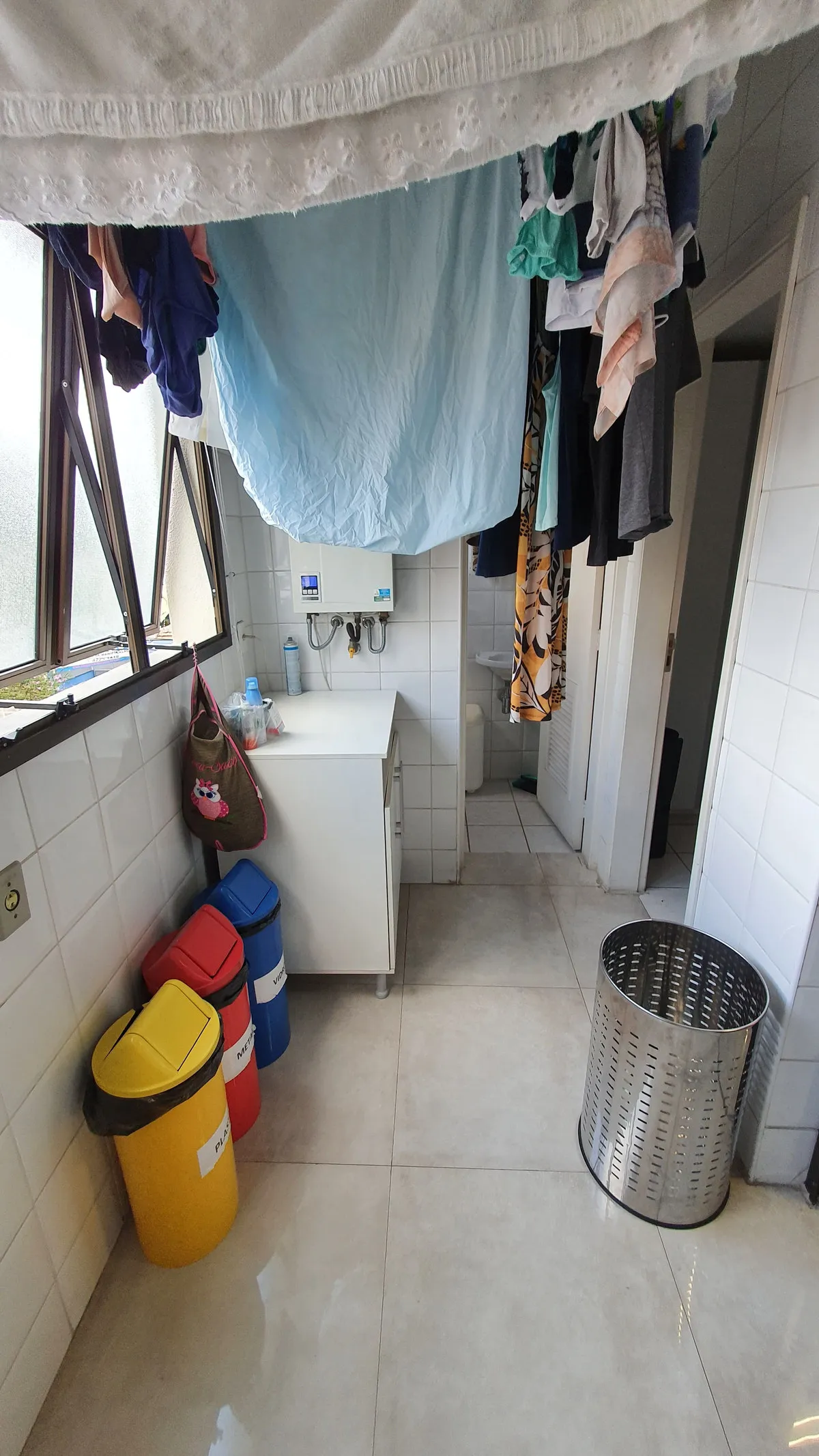 Área de serviço com varal de roupas, aquecedor, lixeiras coloridas e azulejo branco, antes da reforma