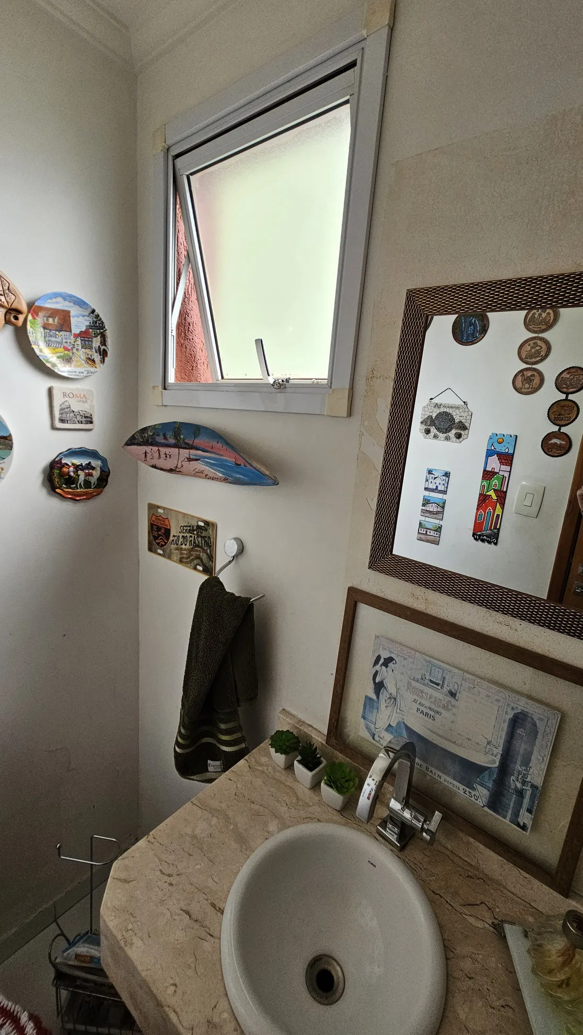 Lavabo com janela basculante, bancada de travertino, cuba oval e placas de viagem na parede, antes da reforma