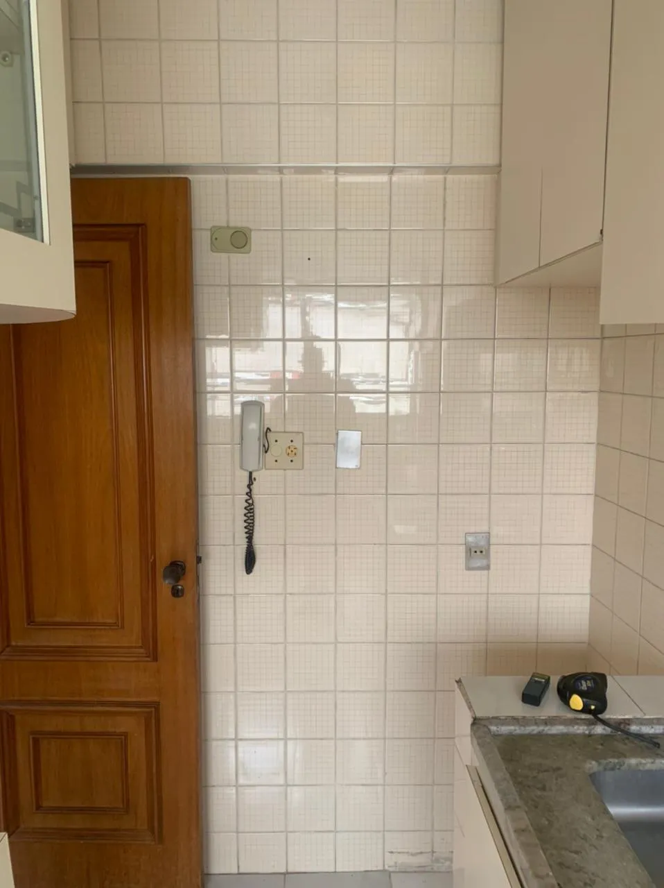 Cozinha com azulejo branco quadriculado, interfone antigo, porta de madeira e bancada de granito com cuba, antes da reforma
