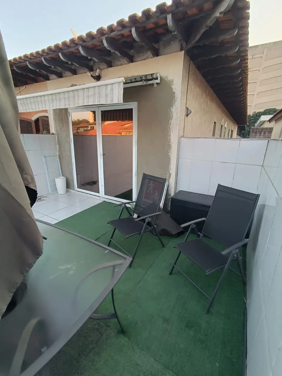 Terraço sob telhado de barro, com grama sintética, cadeiras dobráveis e porta de vidro, antes da reforma