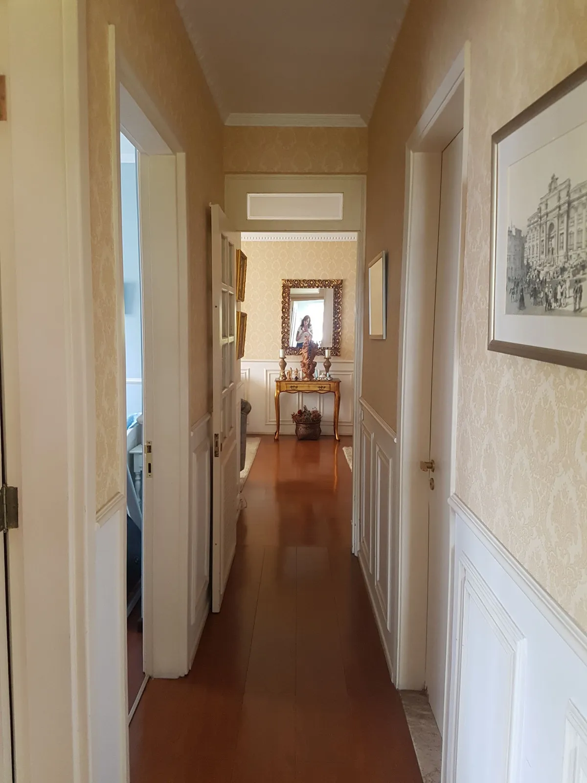 Corredor com papel de parede bege, boiserie branca, piso de madeira alaranjado e aparador ao fundo, antes da reforma