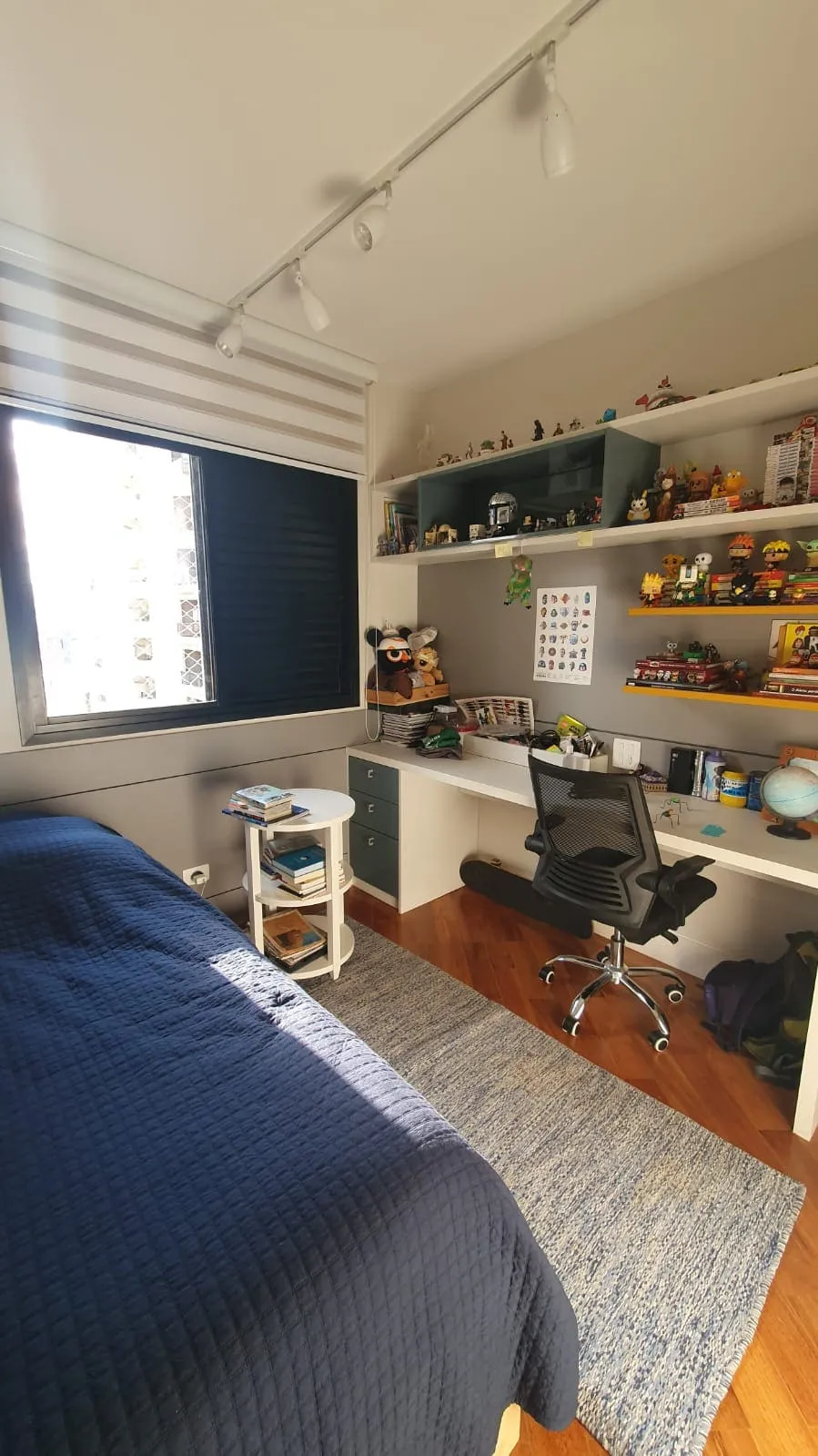 Quarto com cama azul, prateleiras cheias de colecionáveis, escrivaninha branca com cadeira e piso de taco, antes da reforma