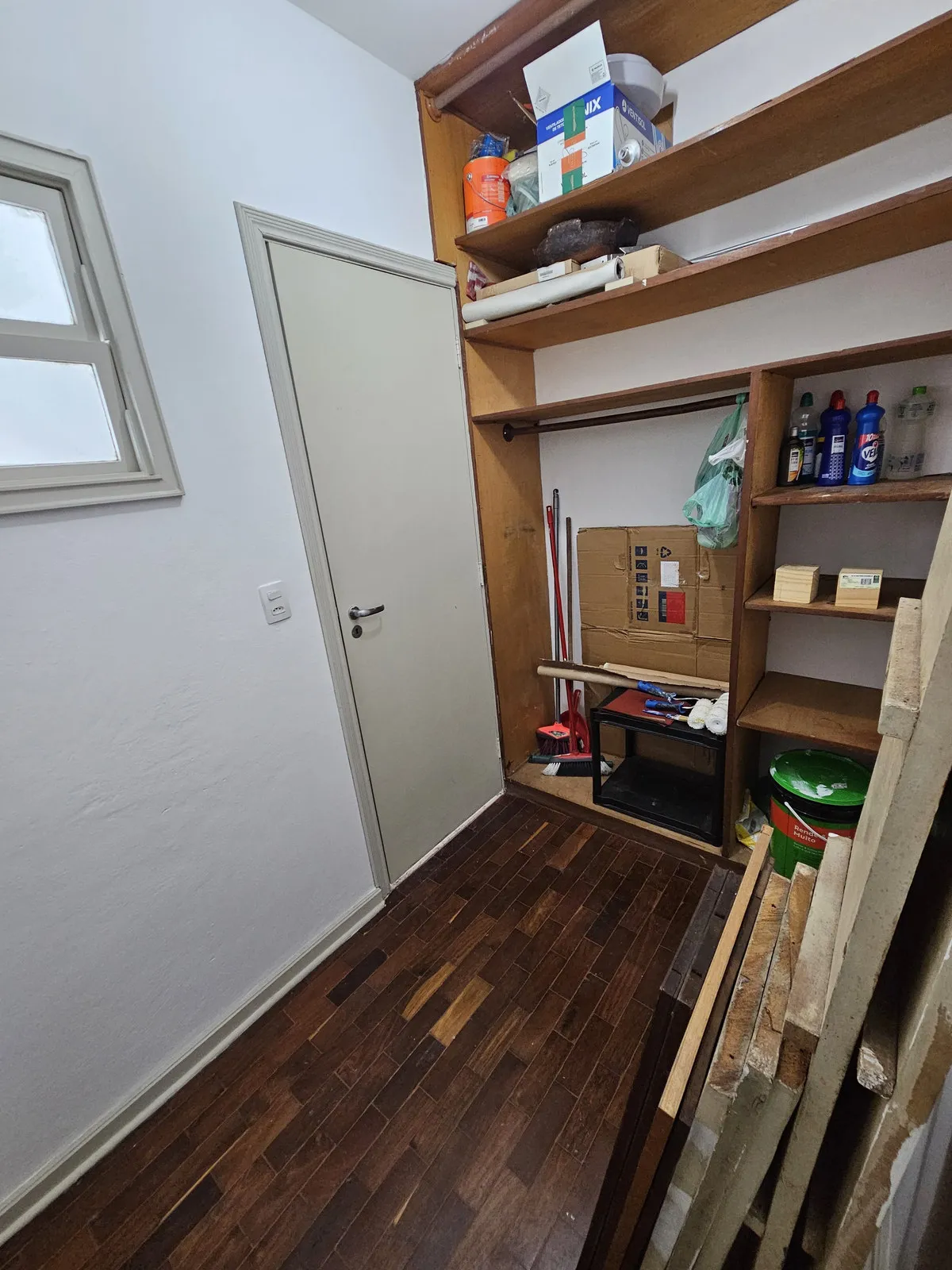 Despensa com prateleiras de madeira, piso de taco escuro e materiais de limpeza guardados, antes da reforma