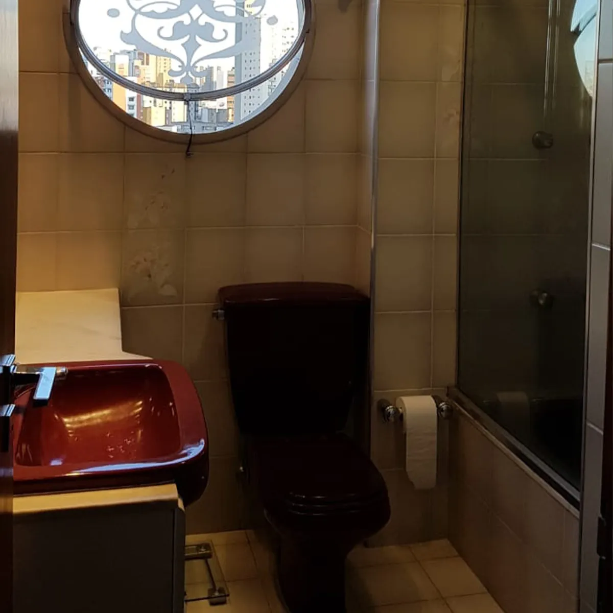 Banheiro antigo com vaso e cuba bordô, azulejo bege, box de vidro e janela redonda, antes da reforma