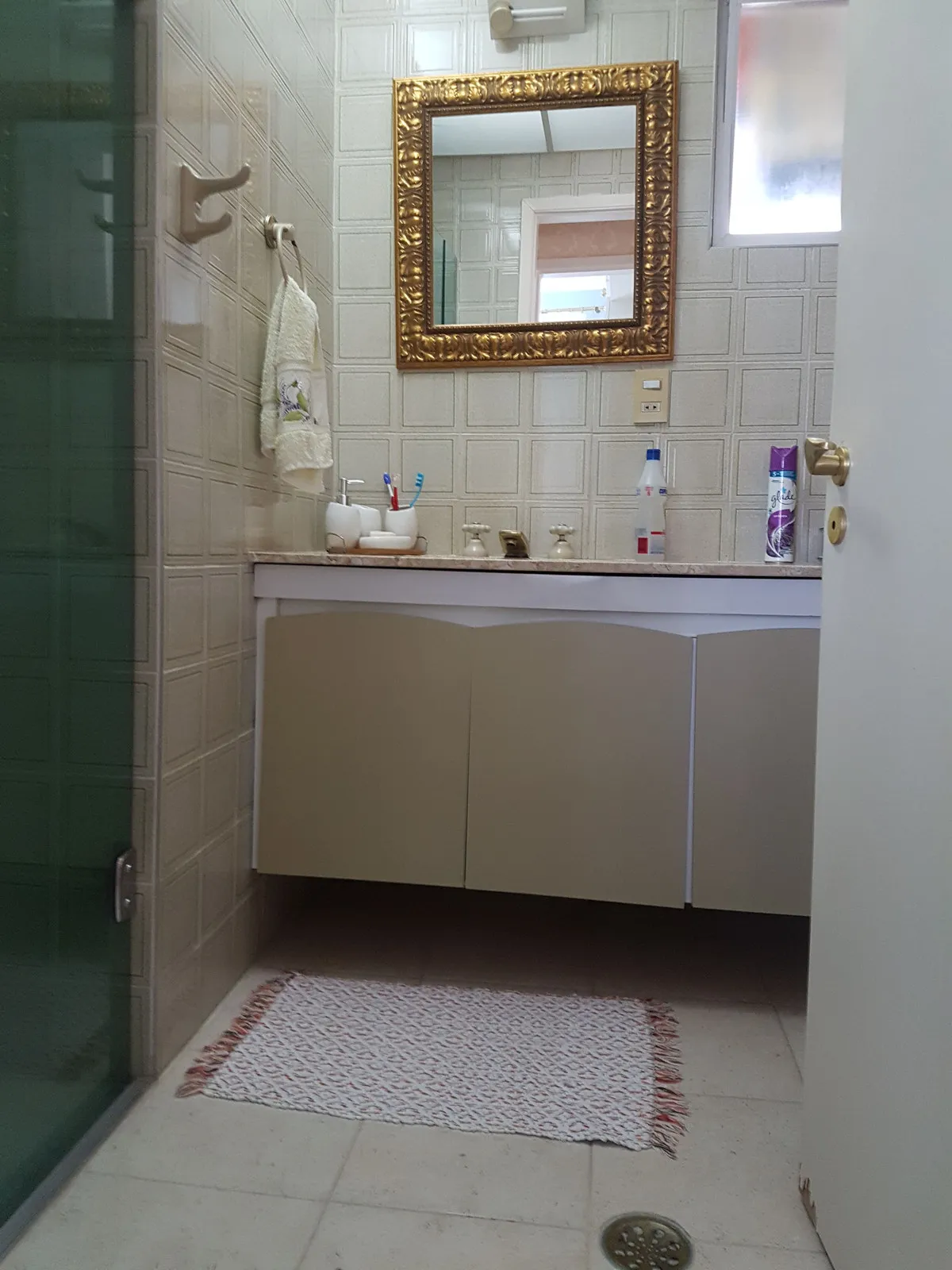 Banheiro com azulejo bege texturizado, gabinete off-white, espelho dourado e box de vidro, antes da reforma