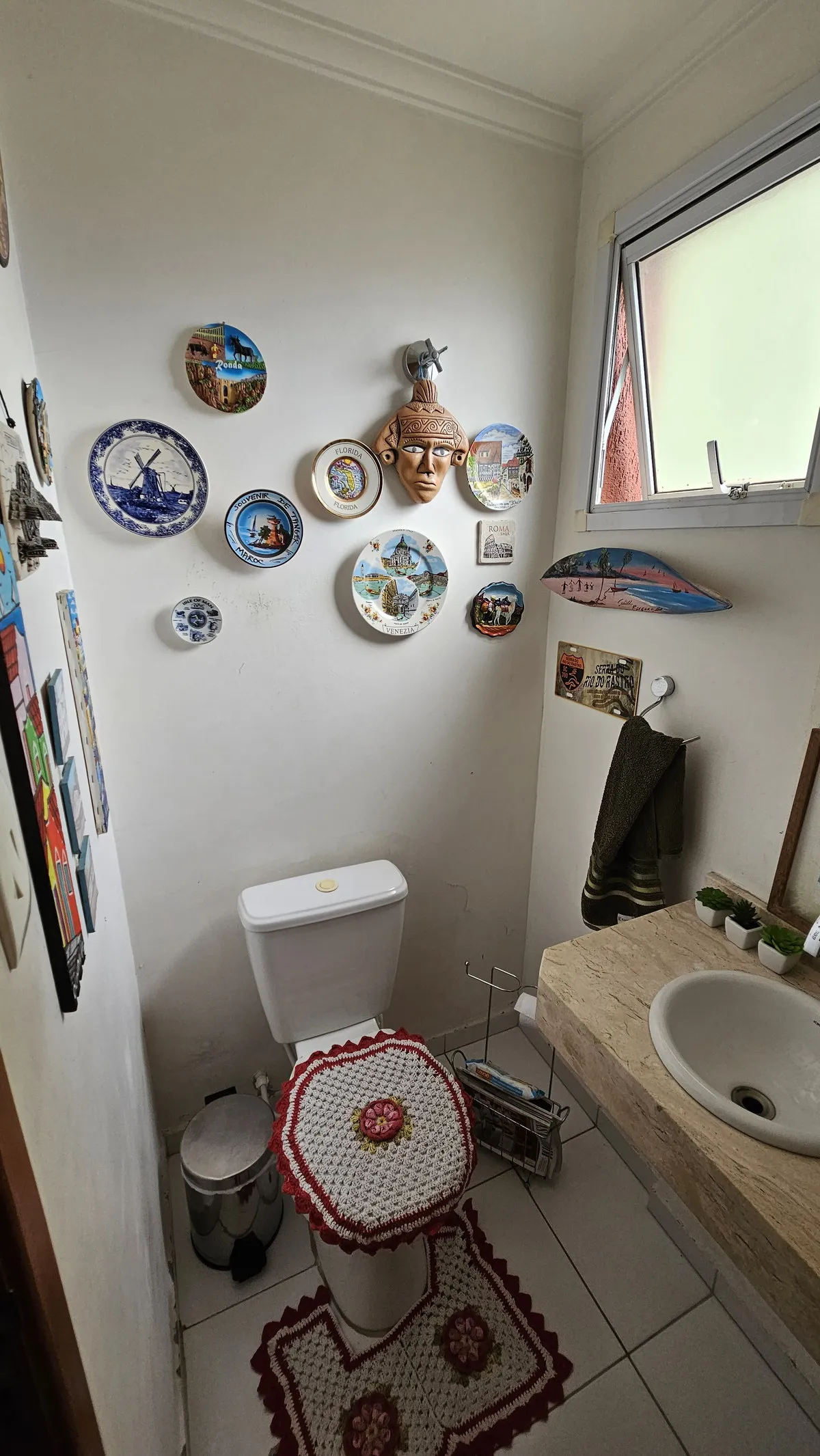 Lavabo com vaso sanitário, tampa e tapete de crochê e parede coberta de pratos de viagem, antes da reforma