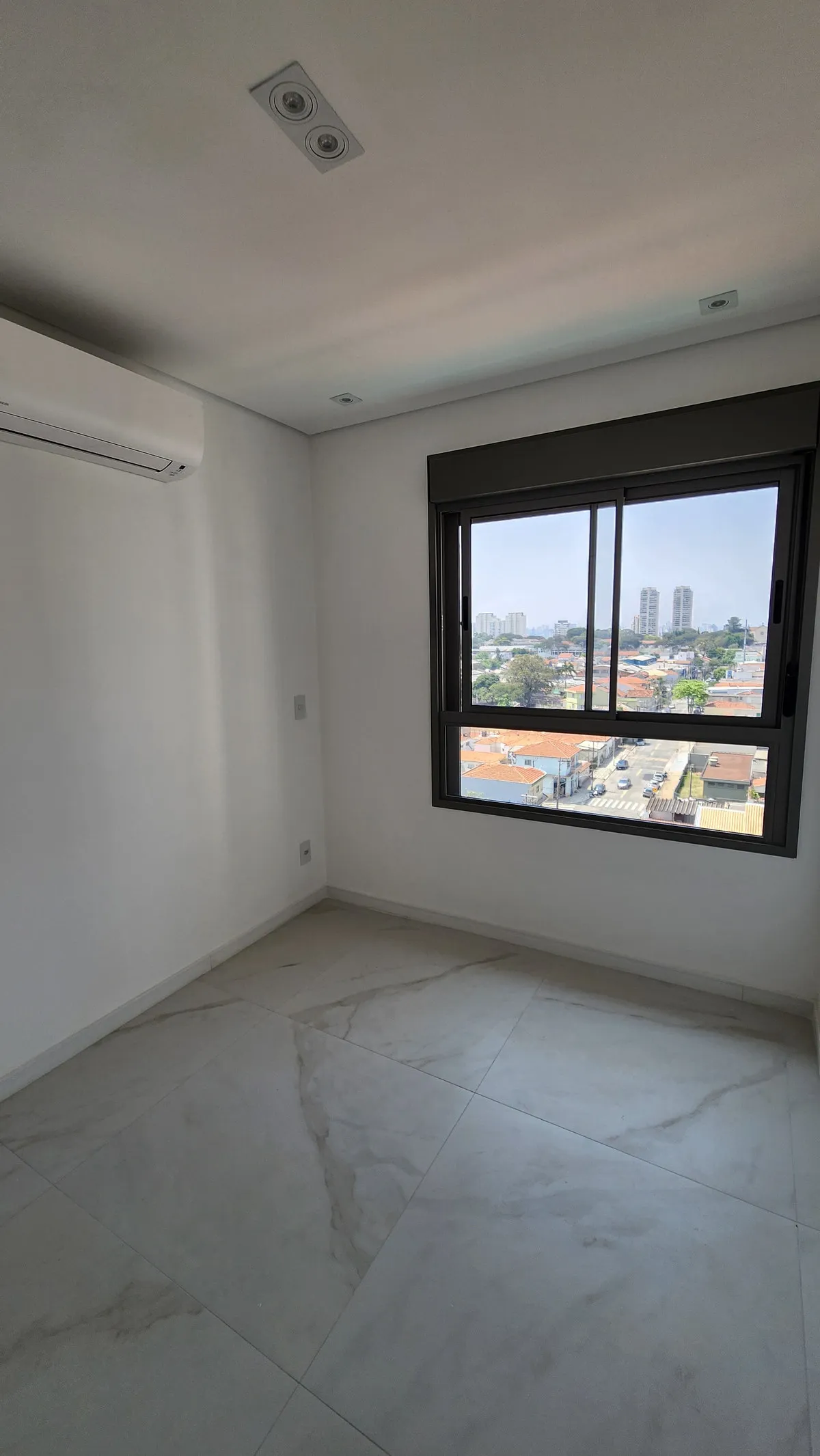 Quarto vazio com janela ampla e vista da cidade, ar-condicionado na parede, antes da reforma