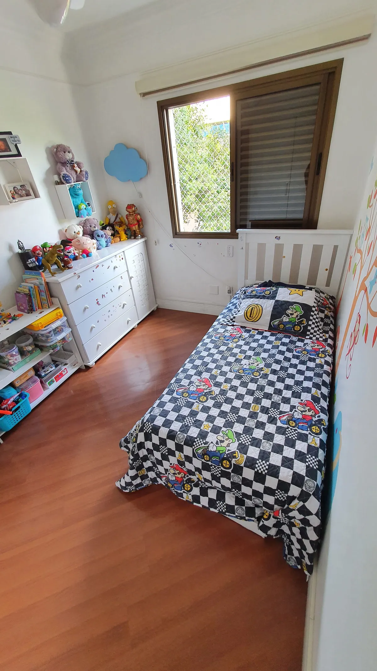 Quarto infantil com colcha do Mario, cômoda branca, brinquedos e piso laminado amadeirado, antes da reforma