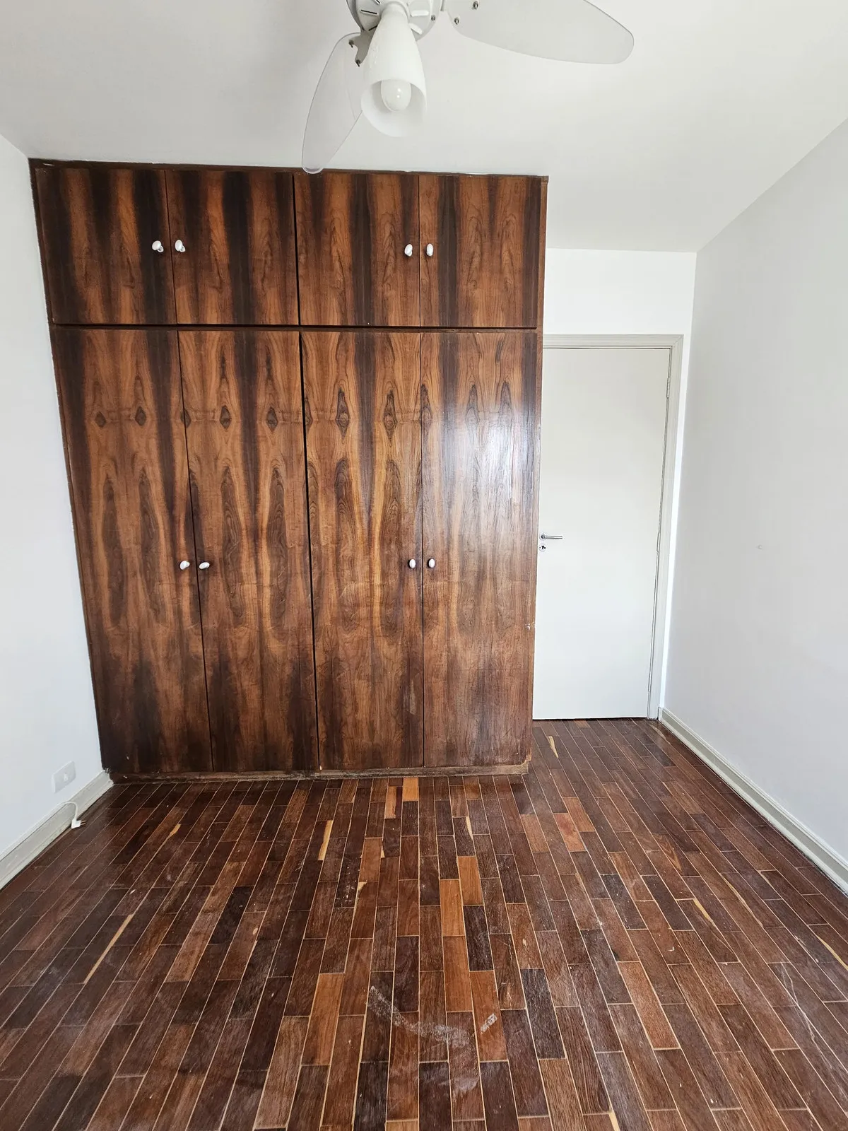 Quarto vazio com guarda-roupa antigo de madeira escura, piso de taco e ventilador de teto, antes da reforma