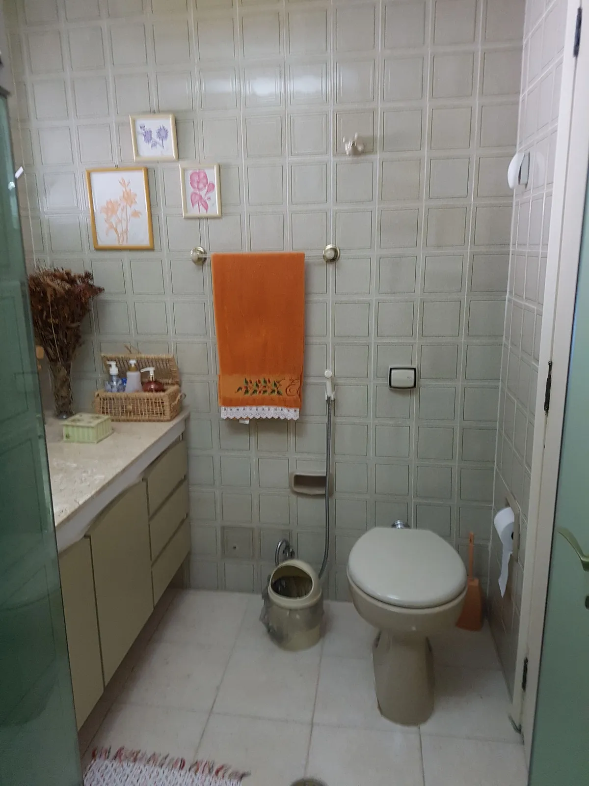 Banheiro com azulejo bege quadriculado, gabinete bege, vaso sanitário antigo e toalha laranja, antes da reforma
