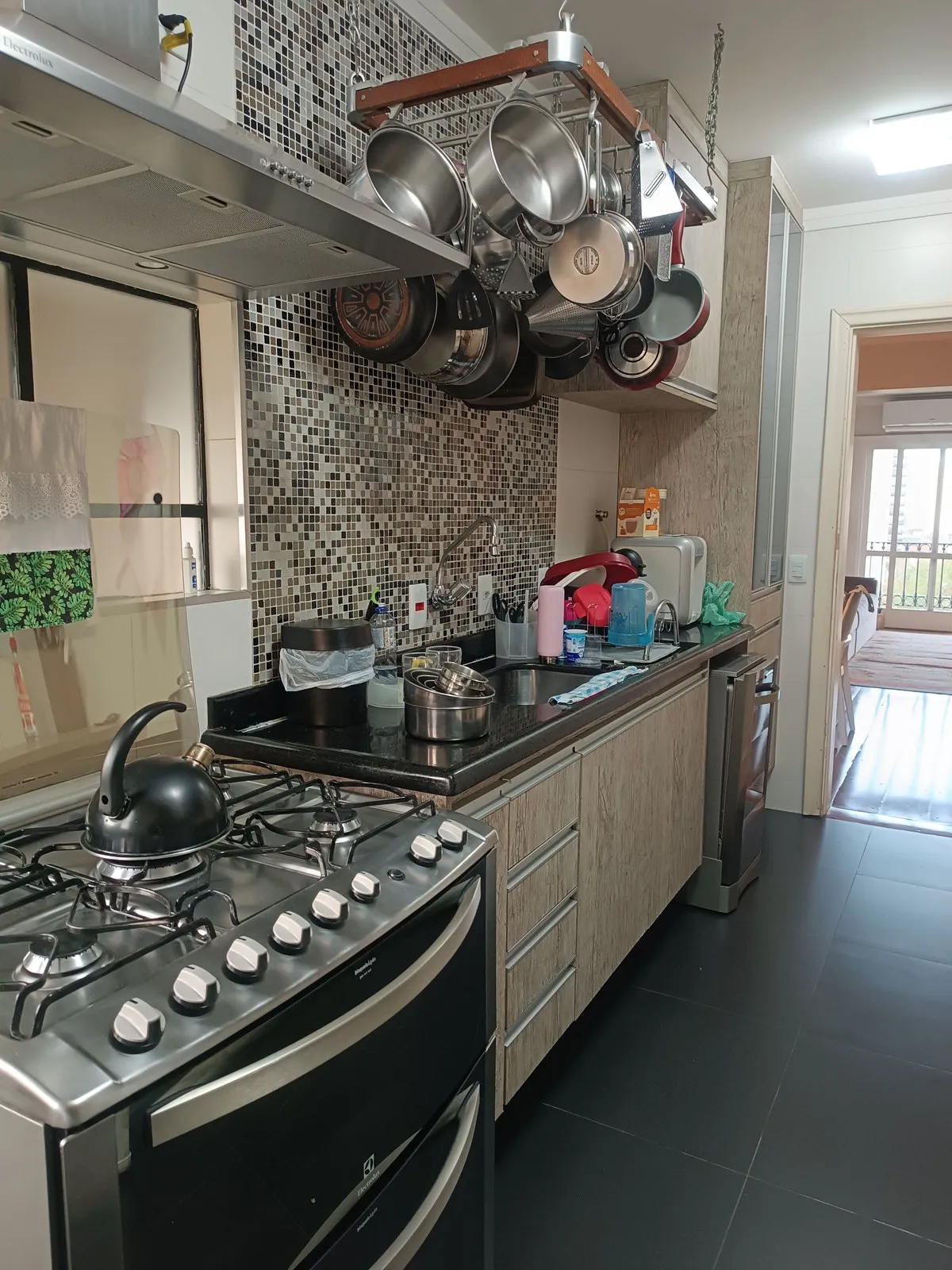 Cozinha corredor com bancada de granito preto, pastilha de vidro, varal de panelas e fogão inox, antes da reforma