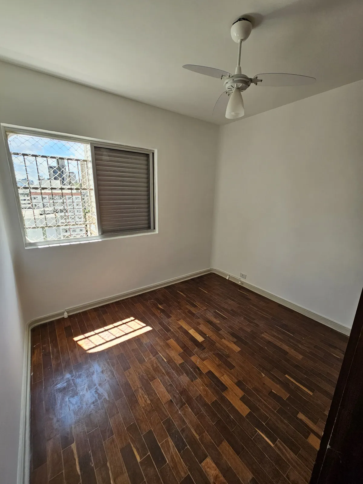 Quarto vazio com paredes brancas, piso de taco escuro e janela com persiana, antes da reforma