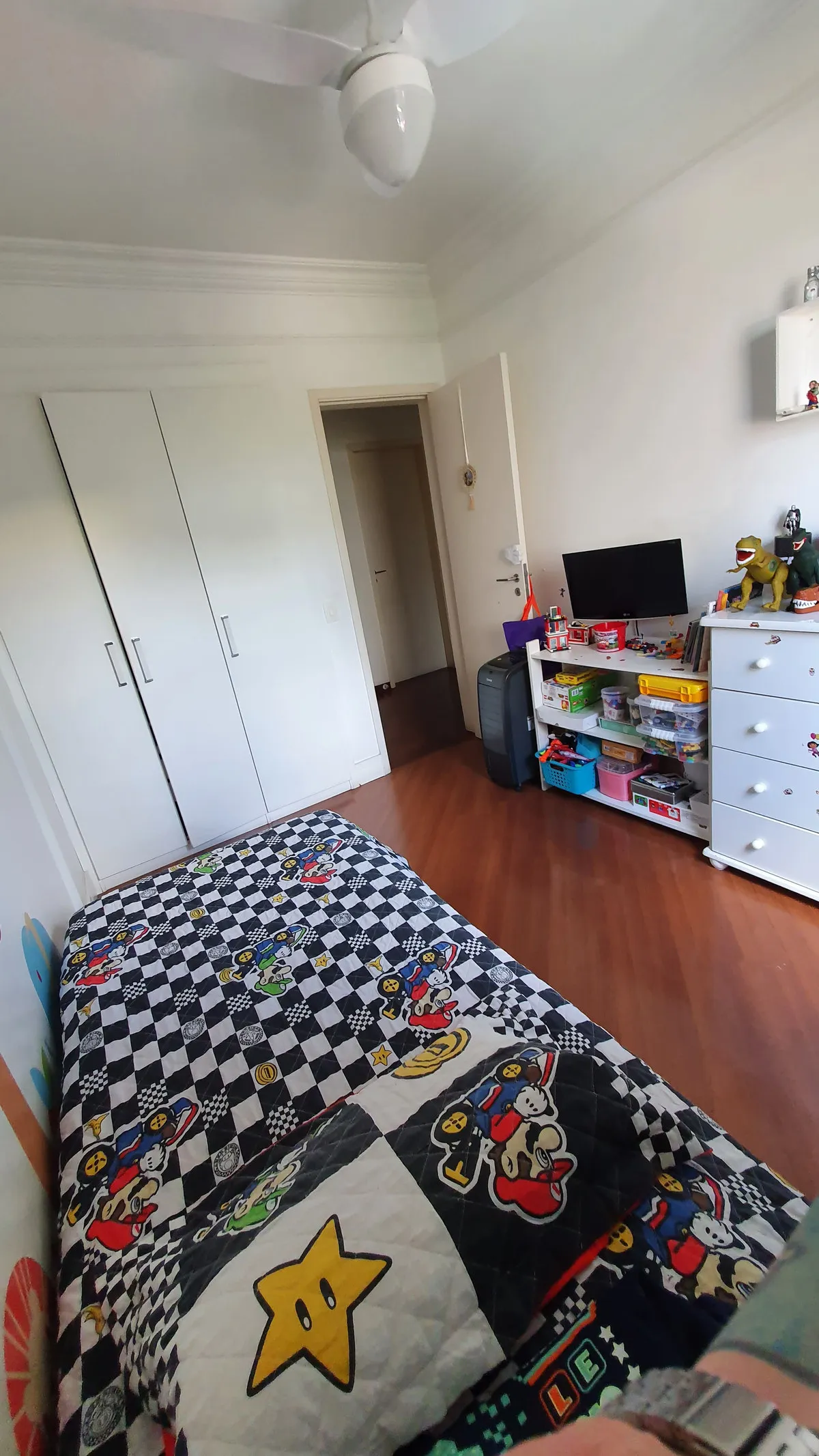 Quarto infantil com guarda-roupa branco, colcha do Mario, TV e prateleira de brinquedos, antes da reforma
