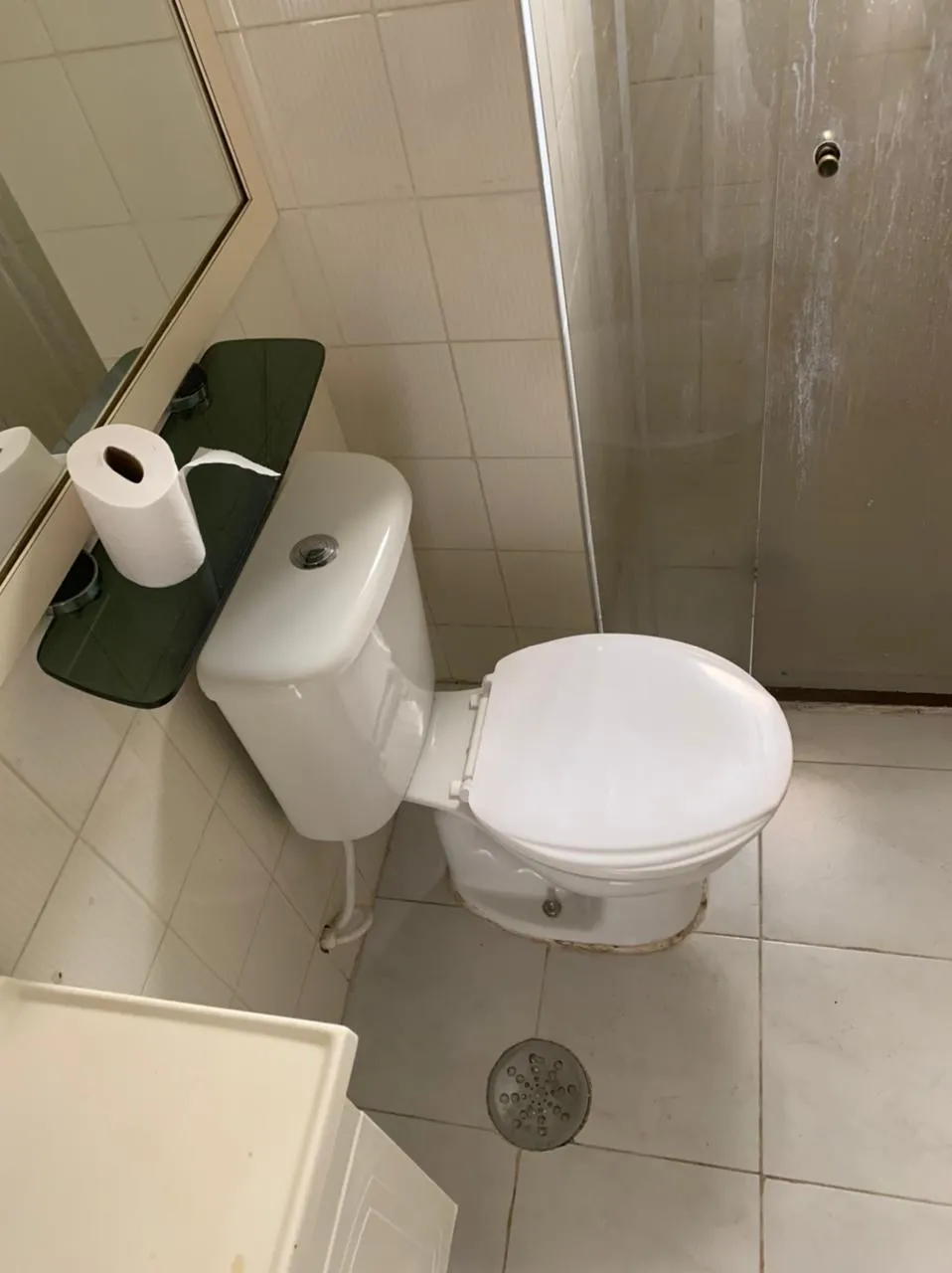 Banheiro com vaso sanitário, box de vidro, azulejo branco e prateleira de vidro verde, antes da reforma
