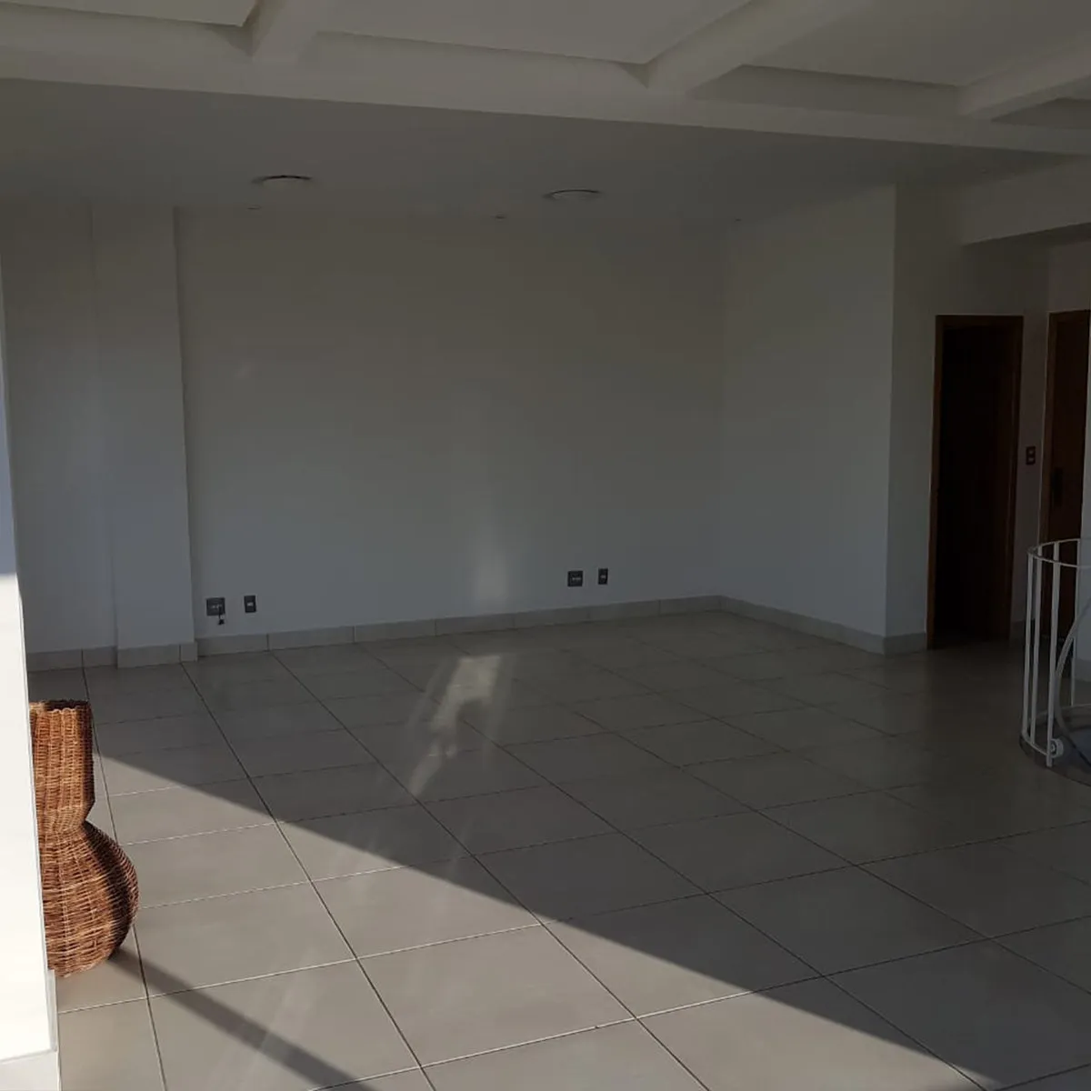 Salão vazio com paredes brancas, piso de porcelanato cinza e porta de madeira, antes da reforma