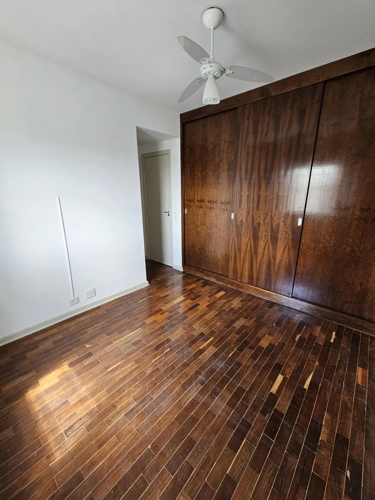 Quarto vazio com guarda-roupa de correr de madeira escura e piso de taco, antes da reforma