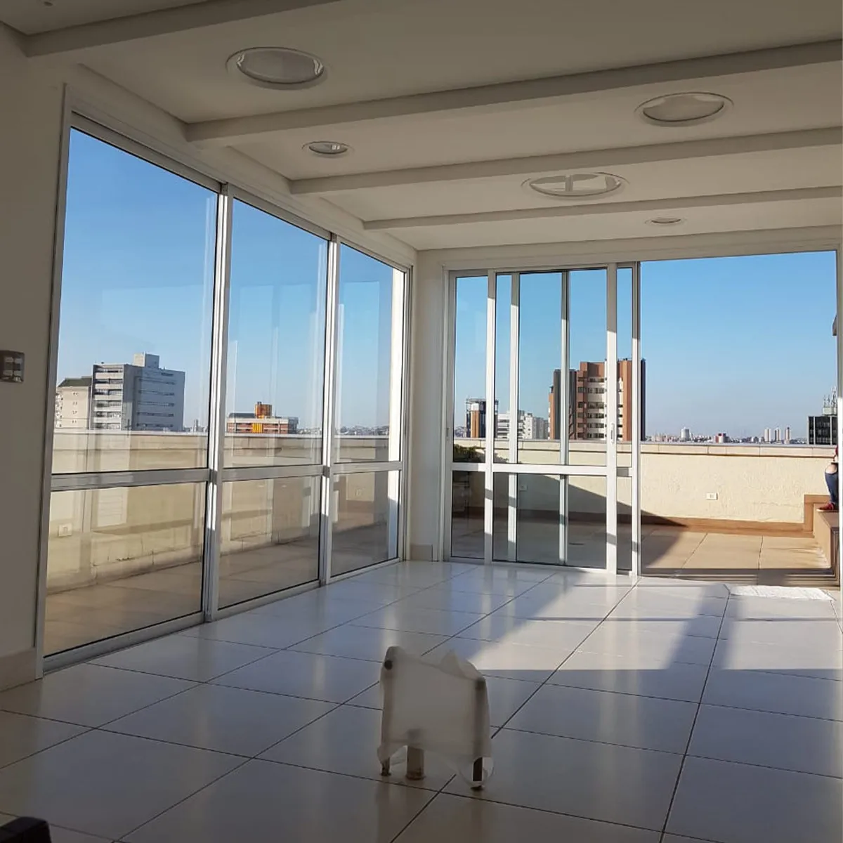 Sala envidraçada vazia na cobertura, com piso branco e vista da cidade, antes da reforma