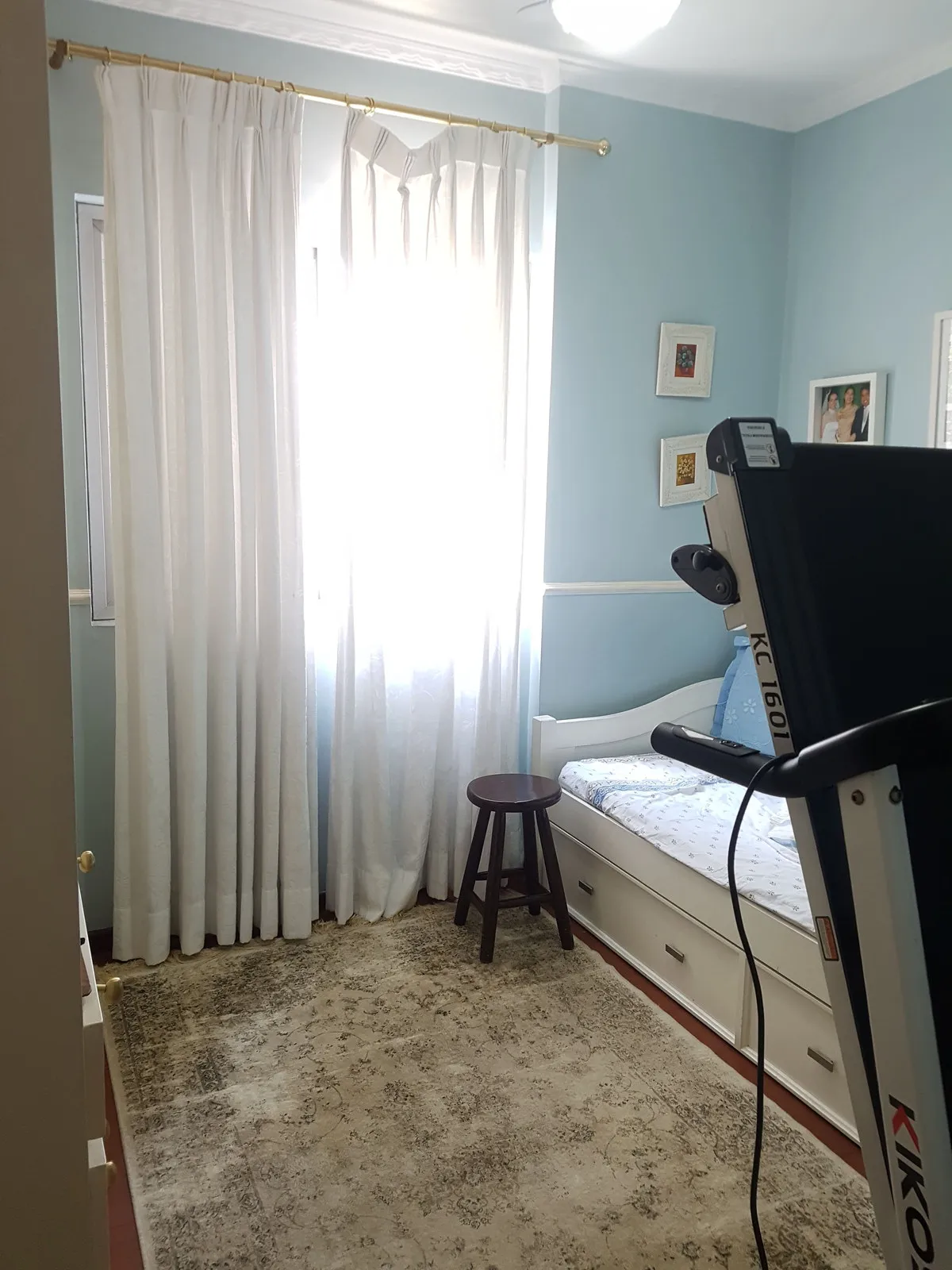 Quarto azul-claro com cama branca, esteira, cortina branca e tapete persa desbotado, antes da reforma