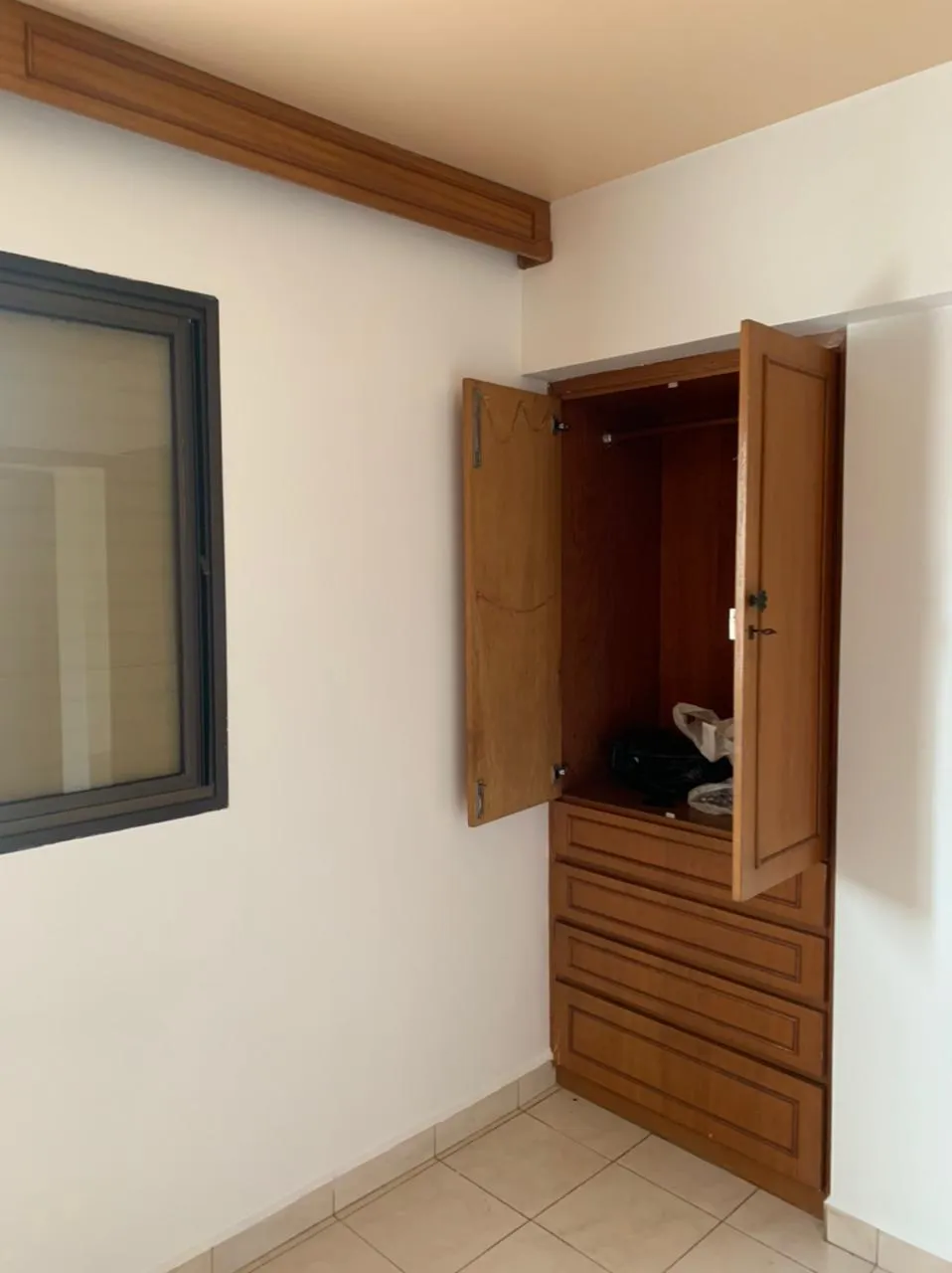 Quarto com parede branca, janela de moldura de madeira e armário embutido de madeira com gavetas, antes da reforma