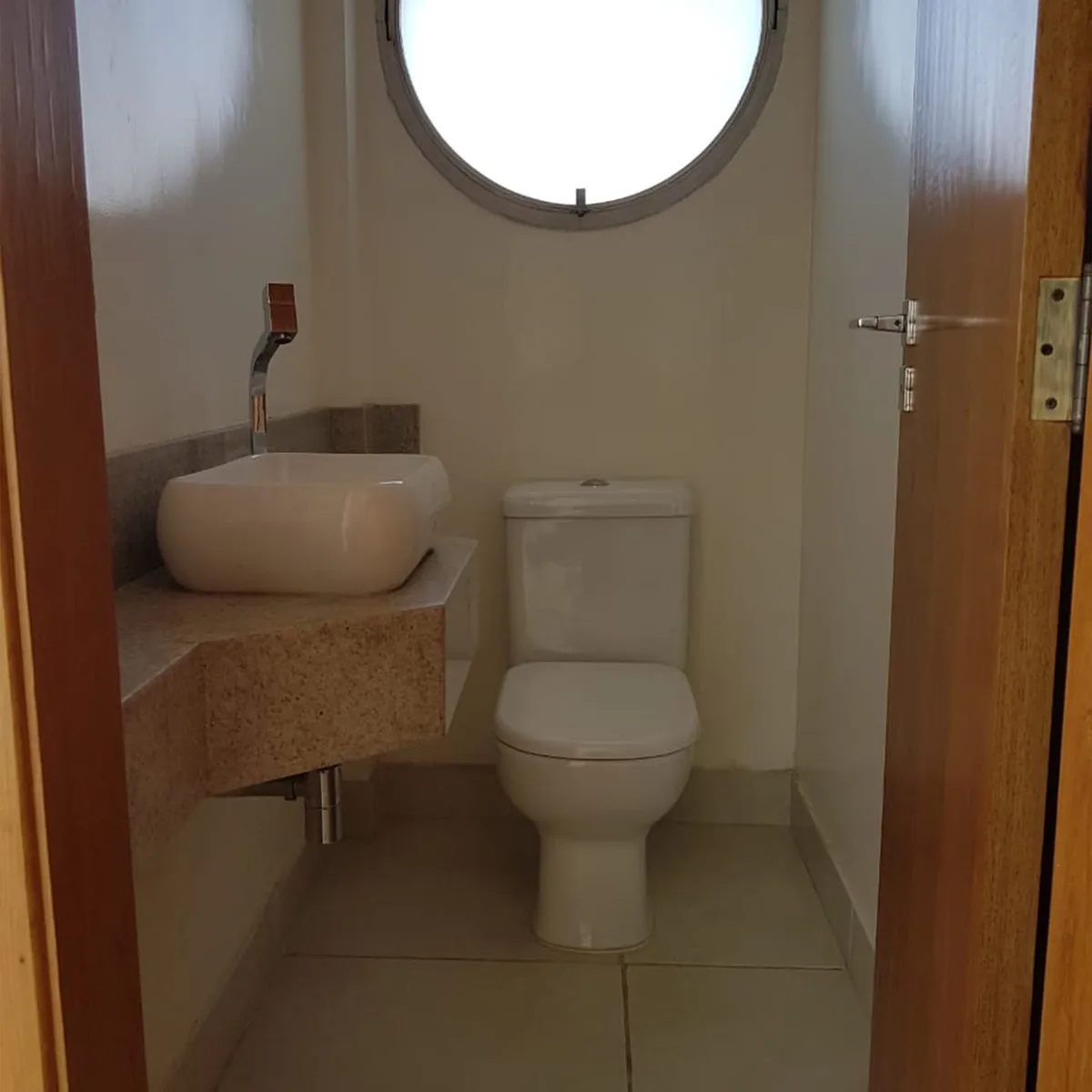 Lavabo antigo com bancada de granito, cuba branca de apoio, vaso e janela redonda, antes da reforma