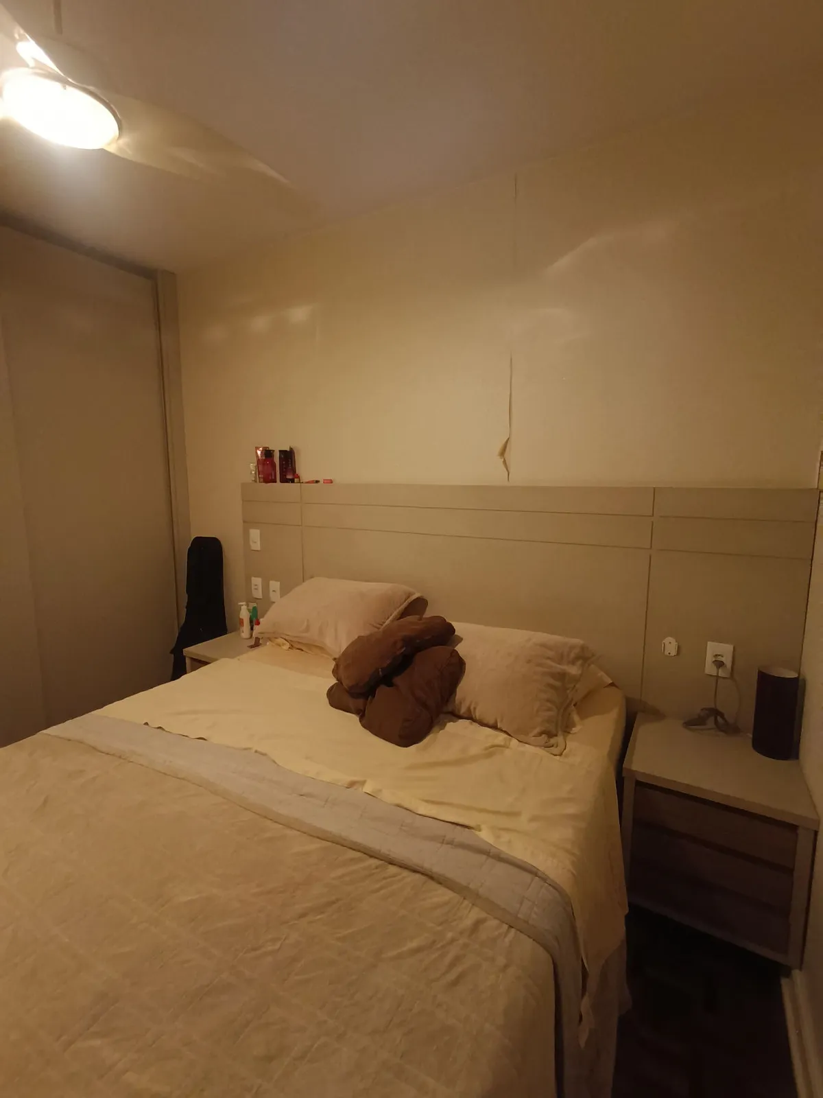 Quarto de casal com cabeceira bege planejada, criado-mudo amadeirado e parede trincada, antes da reforma
