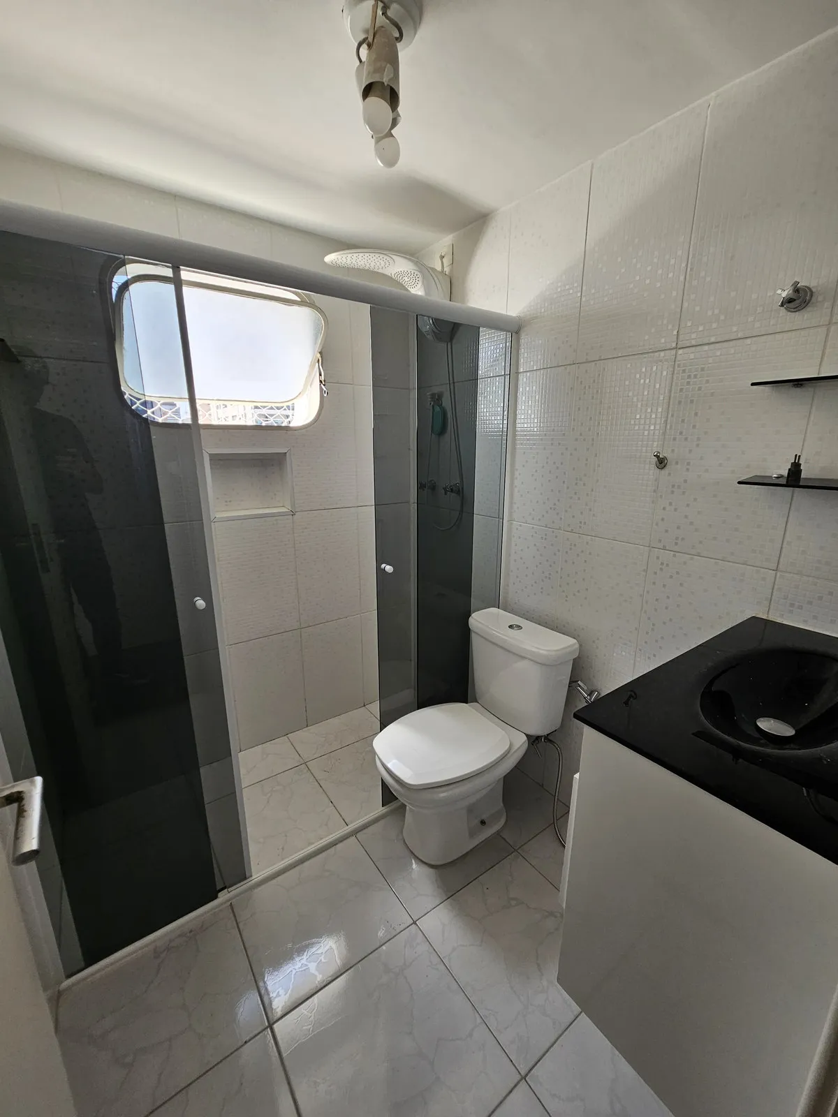 Banheiro com box de vidro fumê, vaso sanitário, cuba preta e azulejo branco, antes da reforma