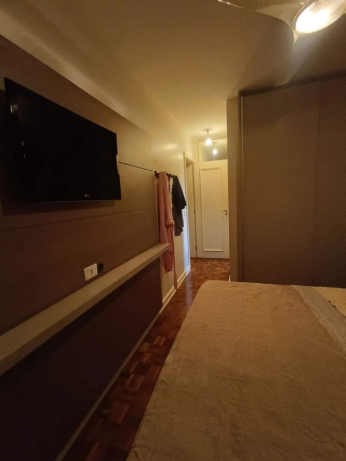 Quarto de casal com painel de madeira, TV, piso de taco e corredor ao fundo, antes da reforma