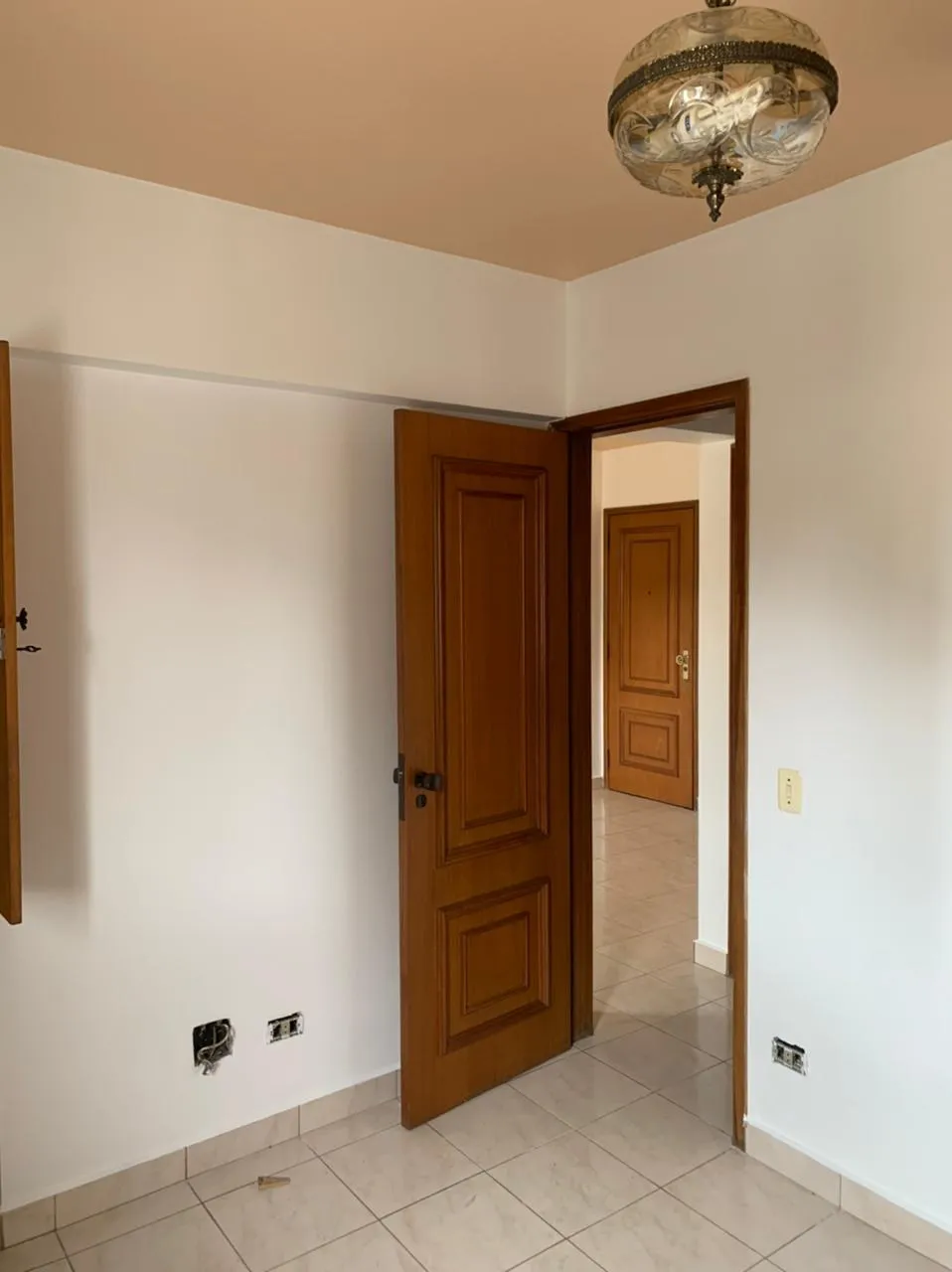 Quarto vazio com teto bege, pendente de vidro, porta de madeira aberta para o corredor e piso de cerâmica, antes da reforma