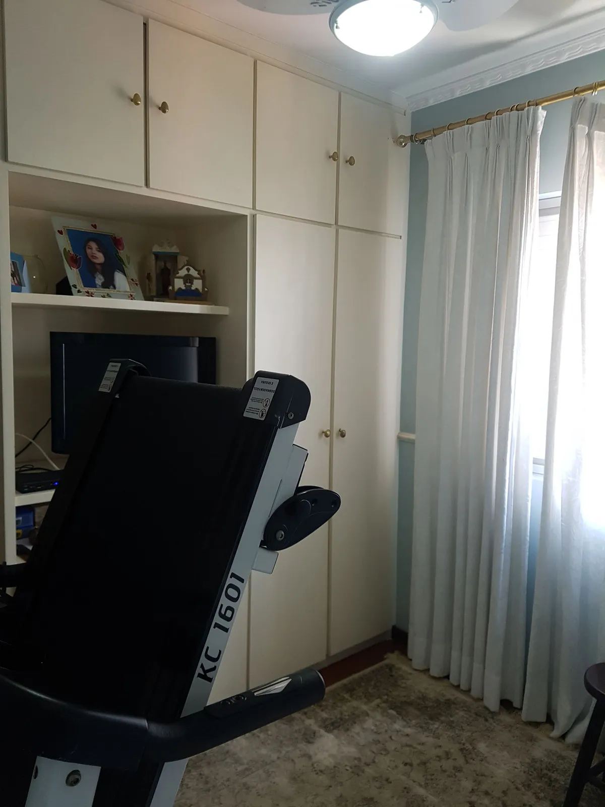 Quarto azul-claro com esteira dobrável, armários brancos embutidos e cortina clara, antes da reforma