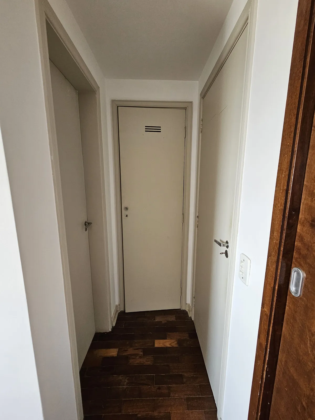Corredor estreito com portas brancas e piso de taco escuro, antes da reforma