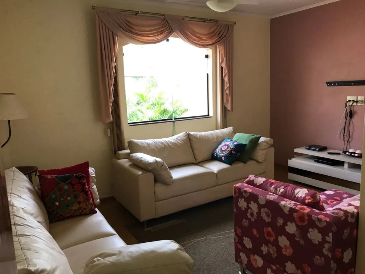 Sala de TV com sofás de couro branco, poltrona floral e cortina de bandô rosa, antes da reforma