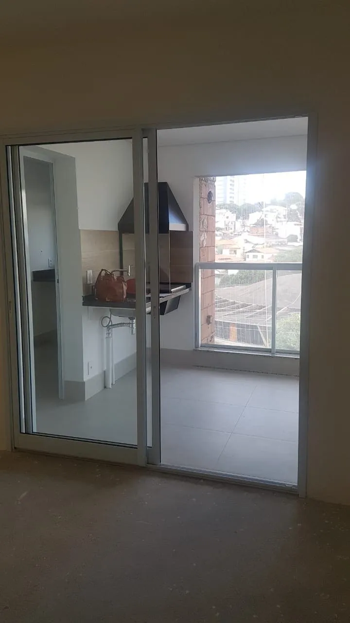 Varanda com churrasqueira e coifa preta vista pela porta de vidro, piso claro e vista da cidade, antes da reforma